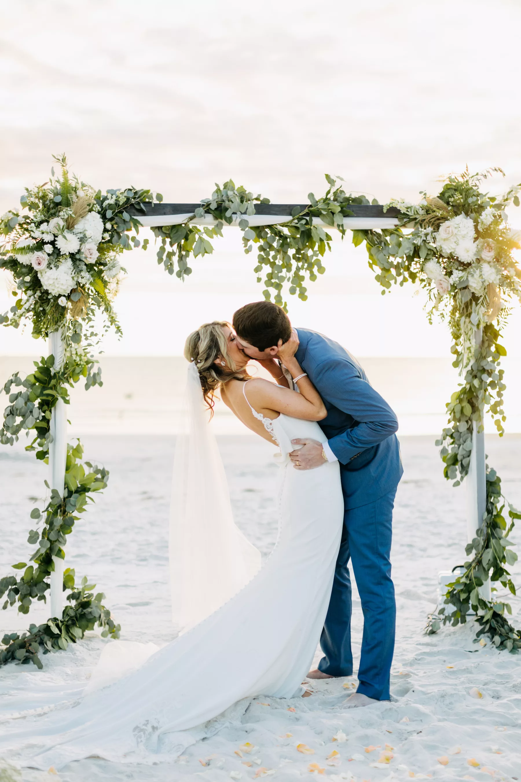 Bride and Groom First Kiss Beach Wedding Portrait | Boho White Arbor with Wrapped Greenery Garland and White Roses | Tampa Bay Videographer Priceless Studio Design | Madeira Beach Photographer Amber McWhorter Photography | Videographer Priceless Studio Design