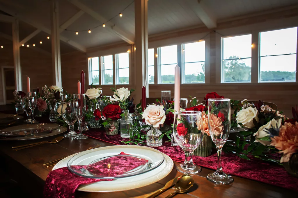 Burgundy Fall Legacy Barn Wedding Reception Inspiration | Rustic Wooden Feasting Table with Red and White Taper Candles, Greenery Garland, Velvet Table Runner, and Bud Vases with Pink and Red Roses, and Orange Chrysanthemums Centerpiece Decor Ideas | Brooksville Florist Save The Date Florida