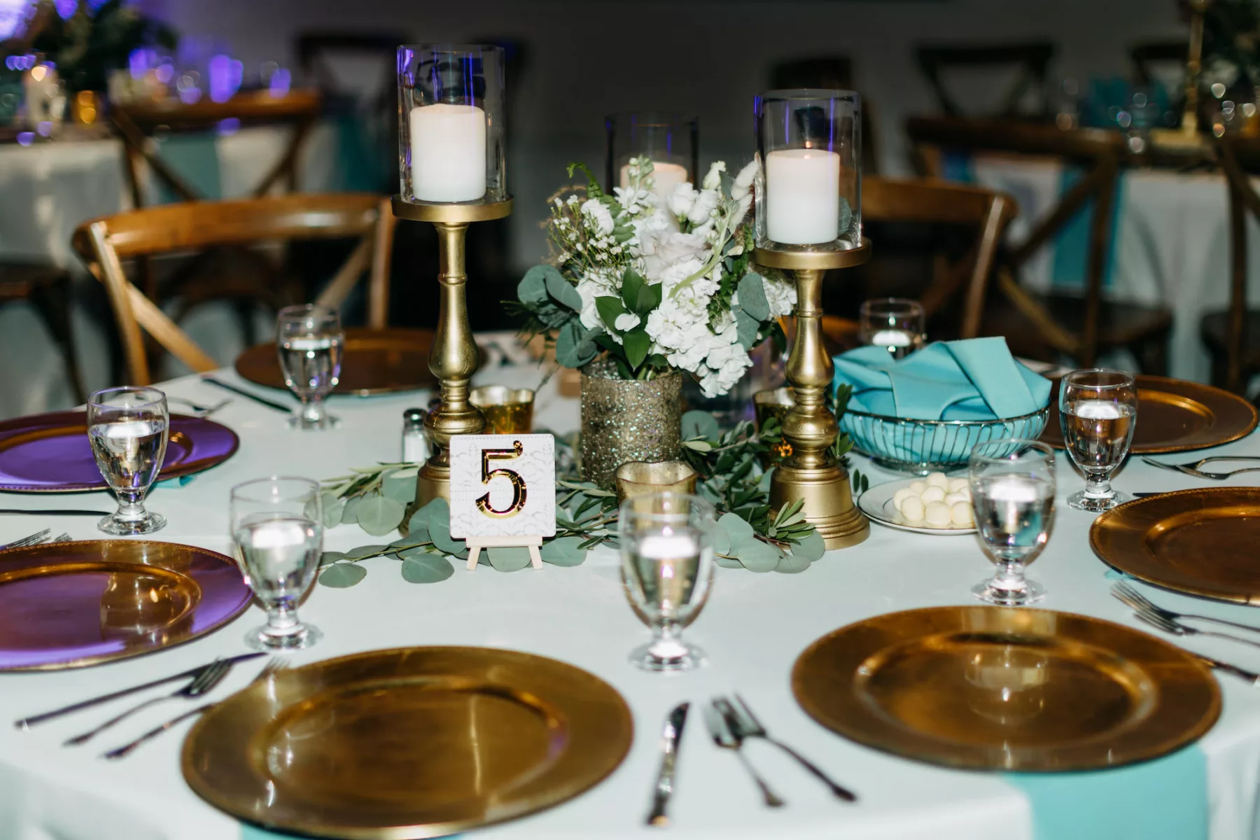 Boho Wedding Reception Centerpiece Decor Ideas | Gold Chargers, Blue Linen Tablescape | Candles, Gold Votives, and Table Numbers | White Flowers, Fern, and Greenery Floral Arrangement Inspiration