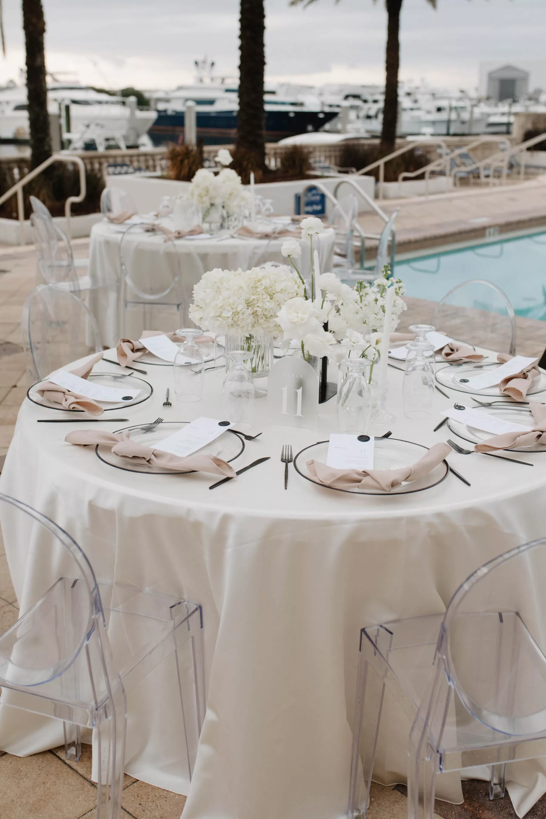 Modern Oceanview Poolside Wedding Reception Floral Centerpiece Decor Ideas | White Hydrangeas, Orchids, and Roses | Frosted Acrylic Table Number Signs | Clear Acrylic Ghost Chairs | Champagne Napkin Linens, Black-Rimmed Chargers, Black Flatware Place Setting Inspiration | Tampa Bay Kate Ryan Event Rentals