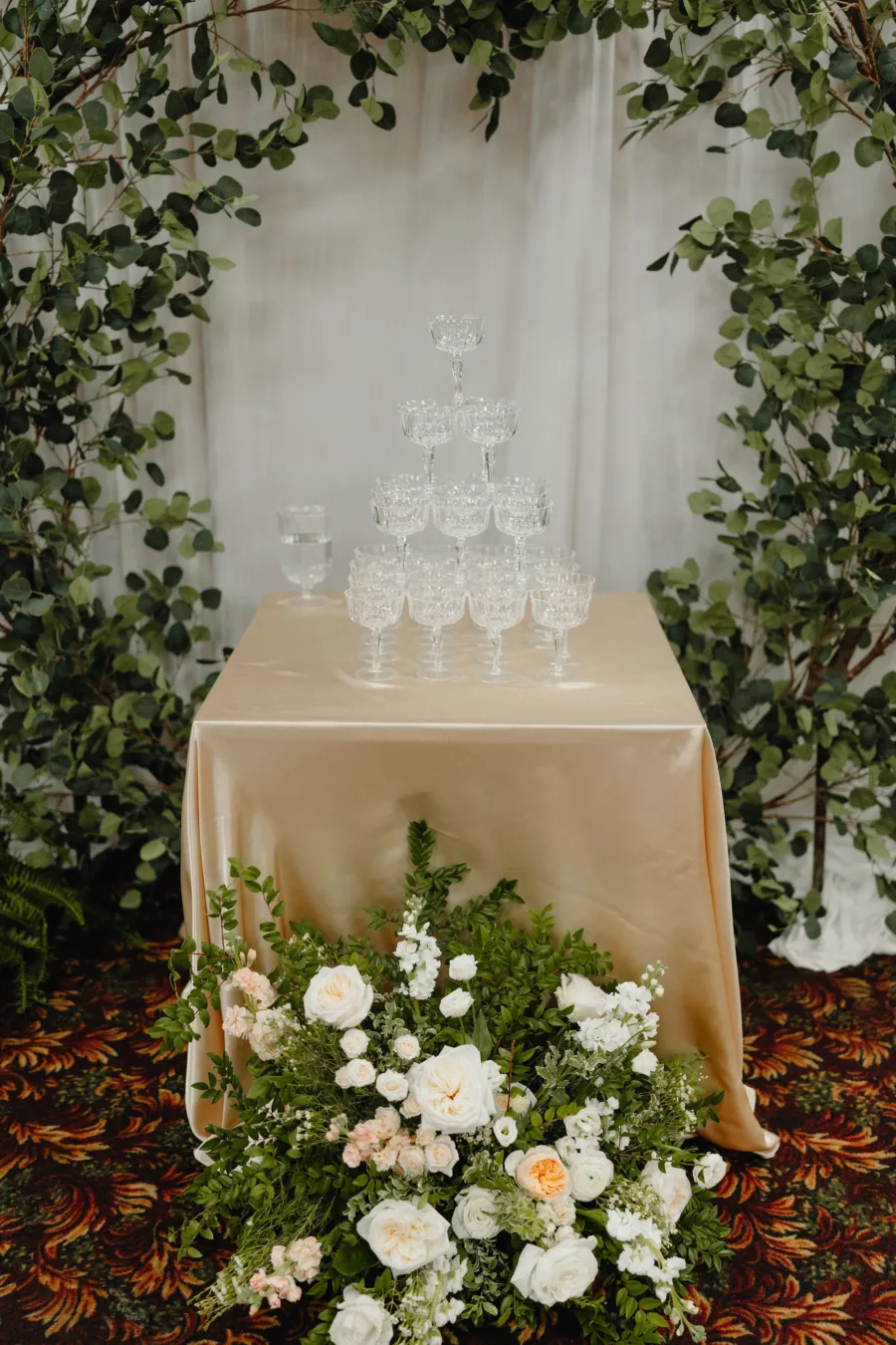 Champagne Tower Table Inspiration with Greenery Backdrop, Champagne Tablecloth, and Italian Inspired Flower Arrangement of Pink and White Roses, and Greenery