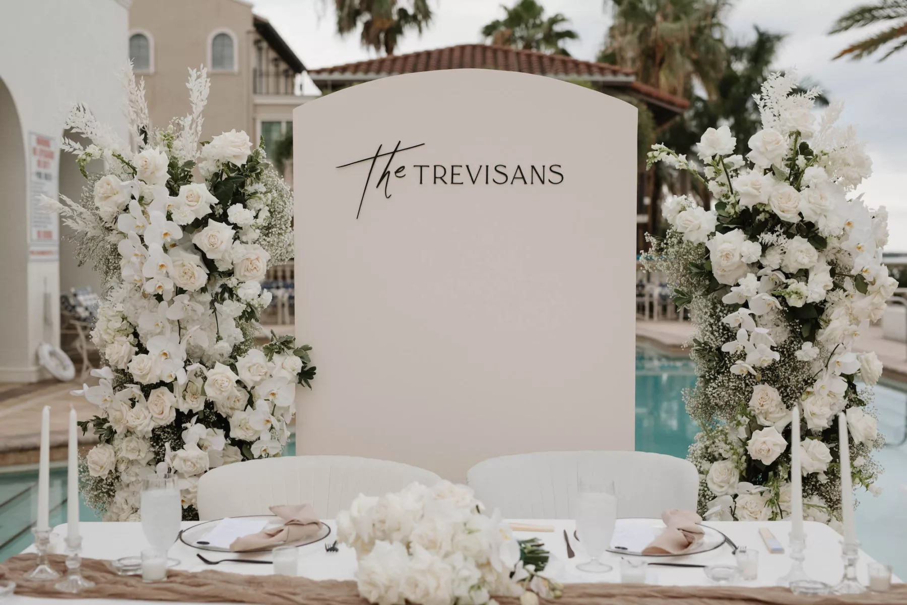 Modern Monochromatic Outdoor Oceanview Spring Wedding Reception Sweetheart Table Decor Inspiration | White Arch Backdrop | Tall Flower Arrangements with White Roses, Carnations, Baby's Breath, and Orchids | White Velvet Chairs | Tampa Bay Kate Ryan Event Rentals