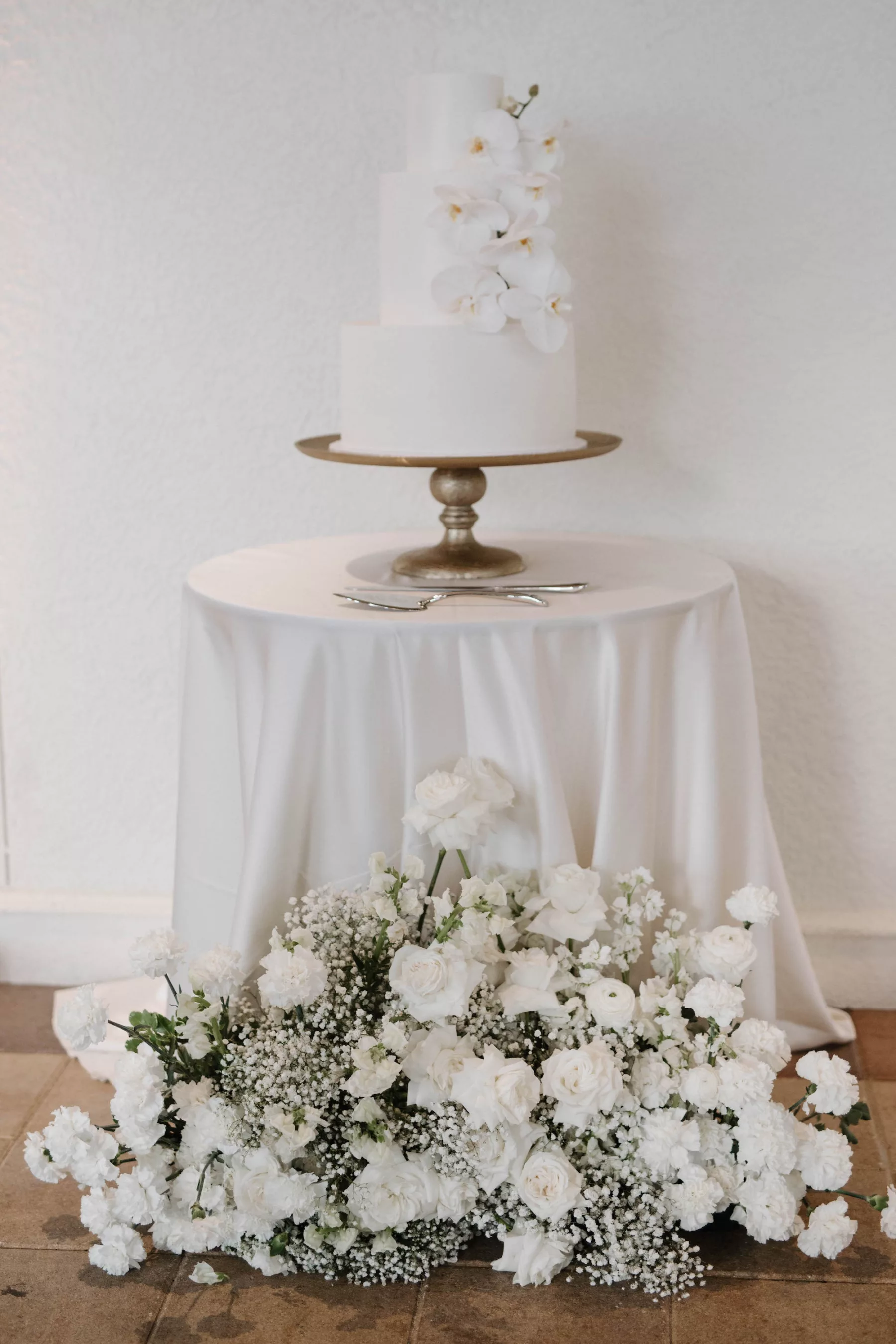 Round Three-Tiered White Wedding Cake with Orchid Accents | Monochromatic Flower Arrangement with White Roses, Baby's Breath, and Carnations | Timeless Cake Table Floral Decor Ideas