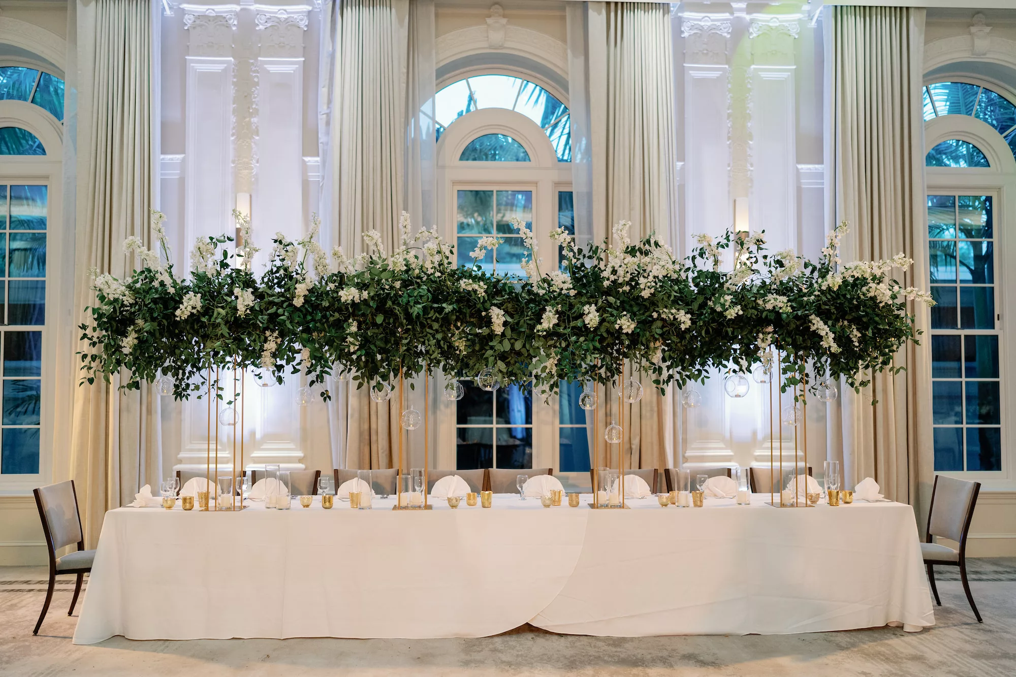 Luxurious White and Gold Grand Ballroom Wedding Reception Head Table Decor Ideas | Hanging Glass Vase Globe Tealight Candle Holder, Long White Feasting Table, Tall Gold Floral Stand with Cascading Greenery and White Flowers | Tampa Bay Event Planner Coastal Coordinating