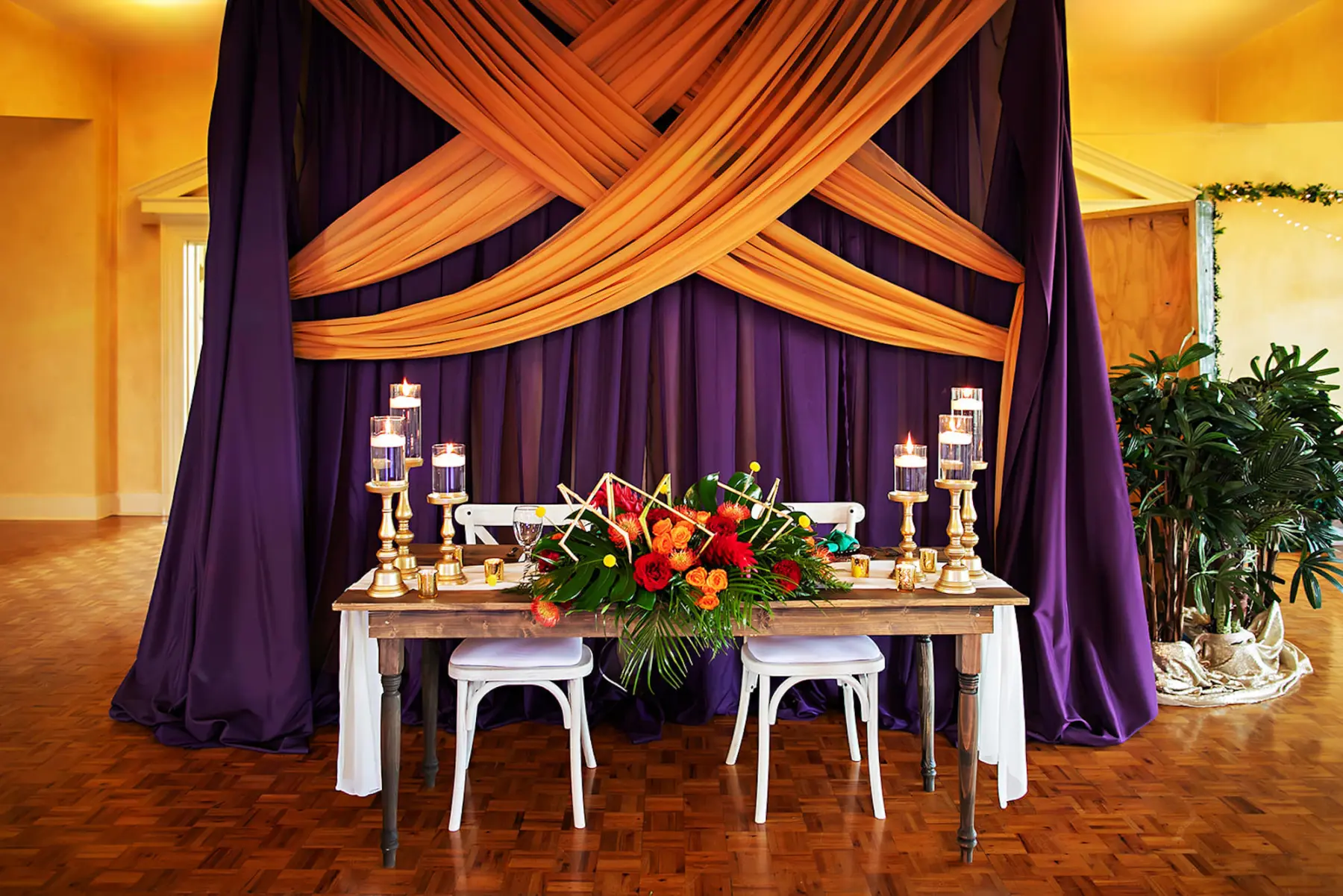 Colorful Geometric Tropical Wedding Reception Sweetheart Table with Orange and Purple Drapery Ideas | Orange Roses and Pincushion Protea, Red Ginger, Carnations, Monstera Leaf, Palm Leaf Floral Decor Inspiration | Tampa Bay Outside The Box Rentals