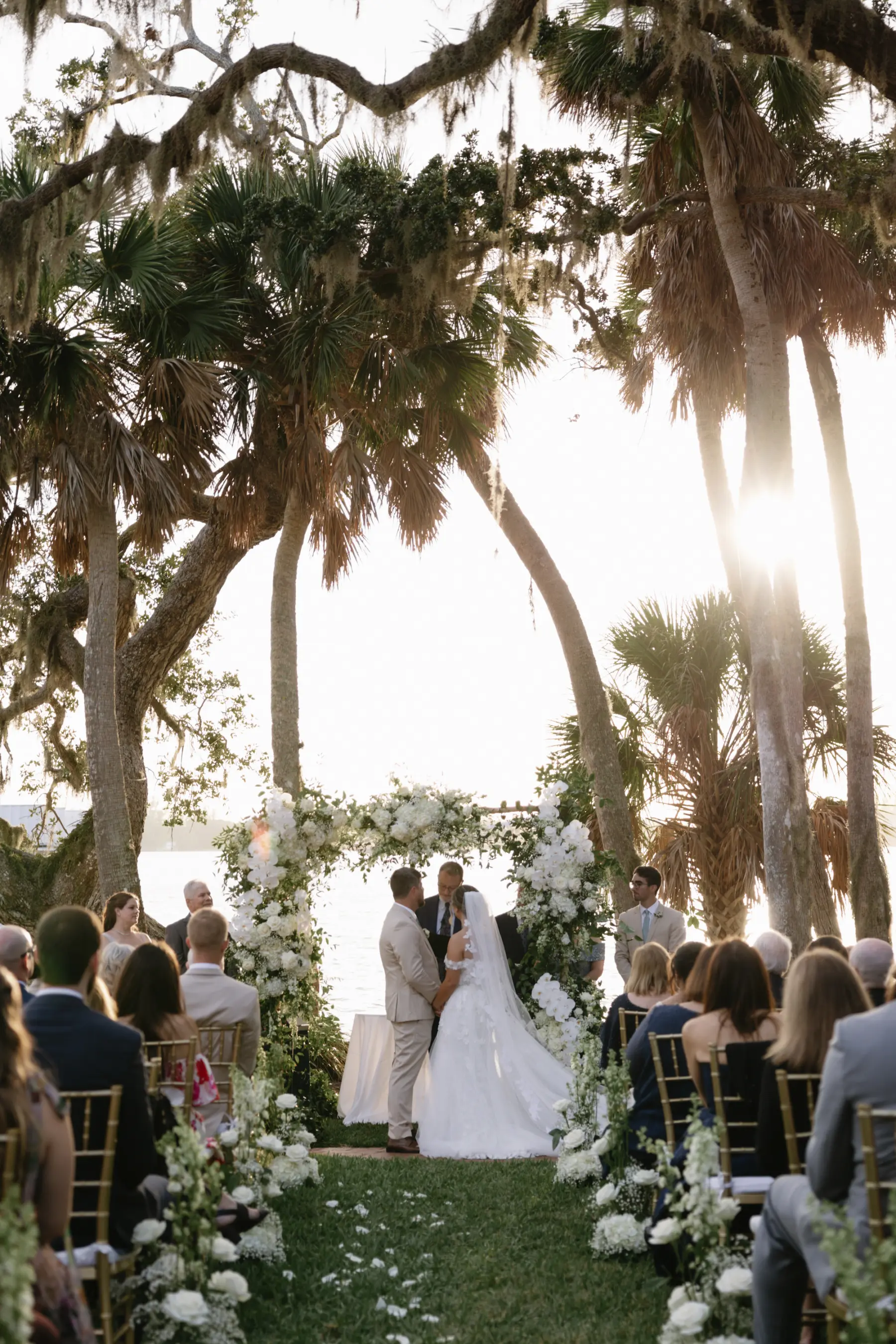 Bride and Groom Outdoor Sunset Oceanfront Wedding Ceremony Ideas | Classic White Wedding Ceremony Arch with White Roses, Orchids, Baby's Breath, Hydrangeas, and Greenery Decor Inspiration