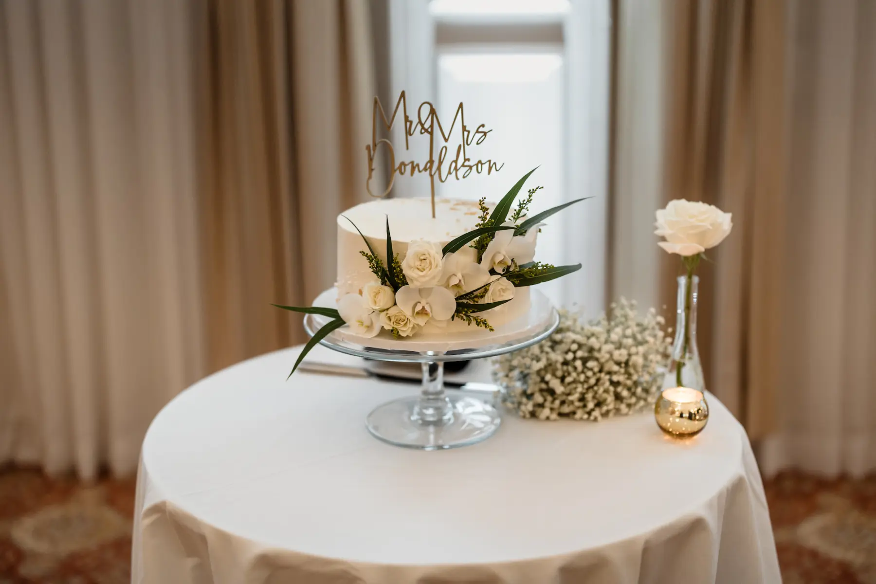 Round White Single Tier Wedding Cake with Custom Cake Topper, White Roses, Orchids, and Greenery Ideas | Classic Cake Table Inspiration