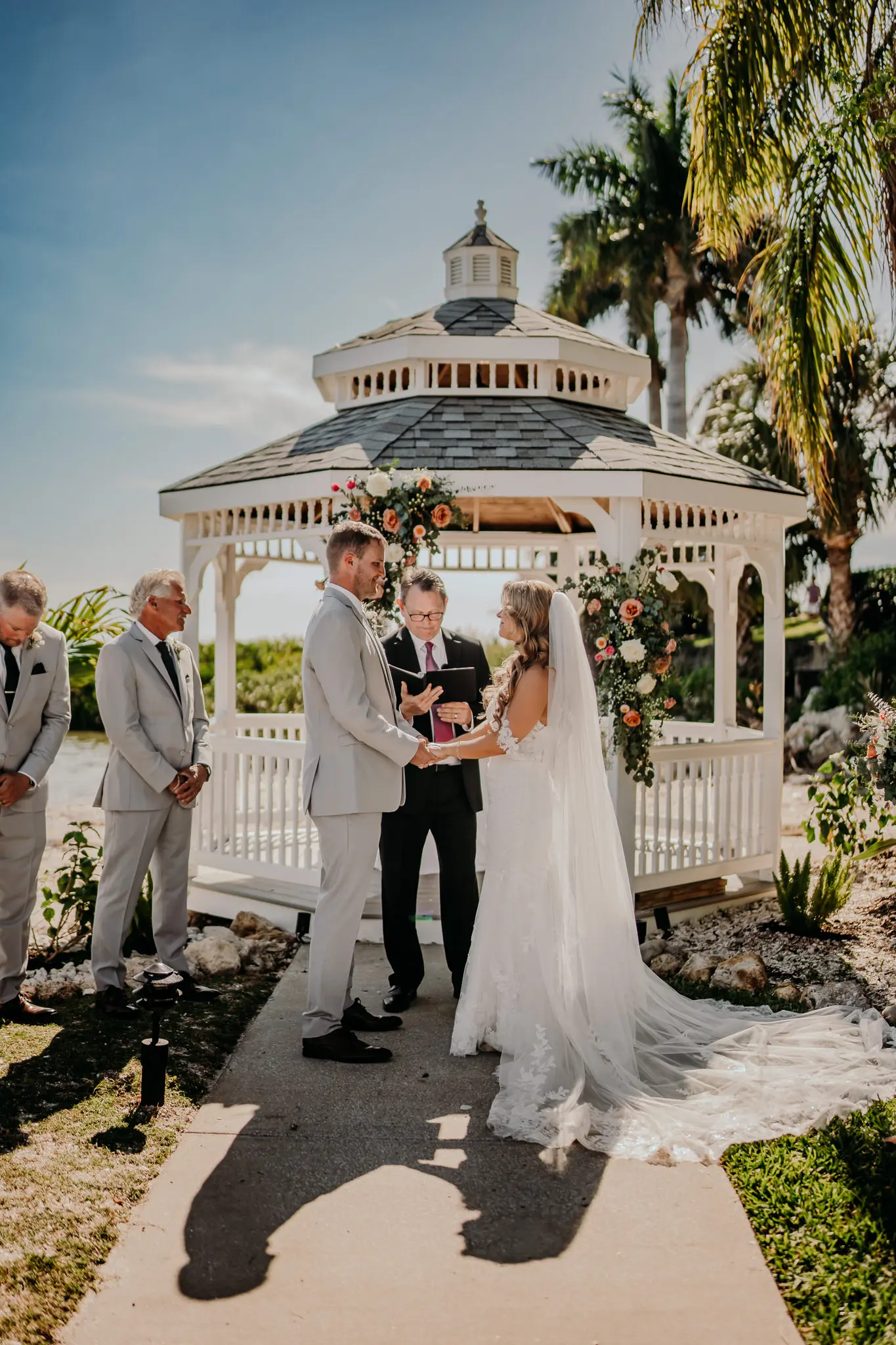 Bride and Groom Outdoor Wedding Ceremony in Front of White Gazebo with Eucalyptus and Soft White and Blush Florals Vow Exchange Wedding | Traditional Long Tulle Veil Inspiration | Groom Gray Suits and Dark Ties Ideas | Off the Shoulder Delicate Lace and Beaded Wedding Gown Inspiration | Tampa Bay Photographer Sabrina Autumn Photography | Tampa Venue Rusty Pelican