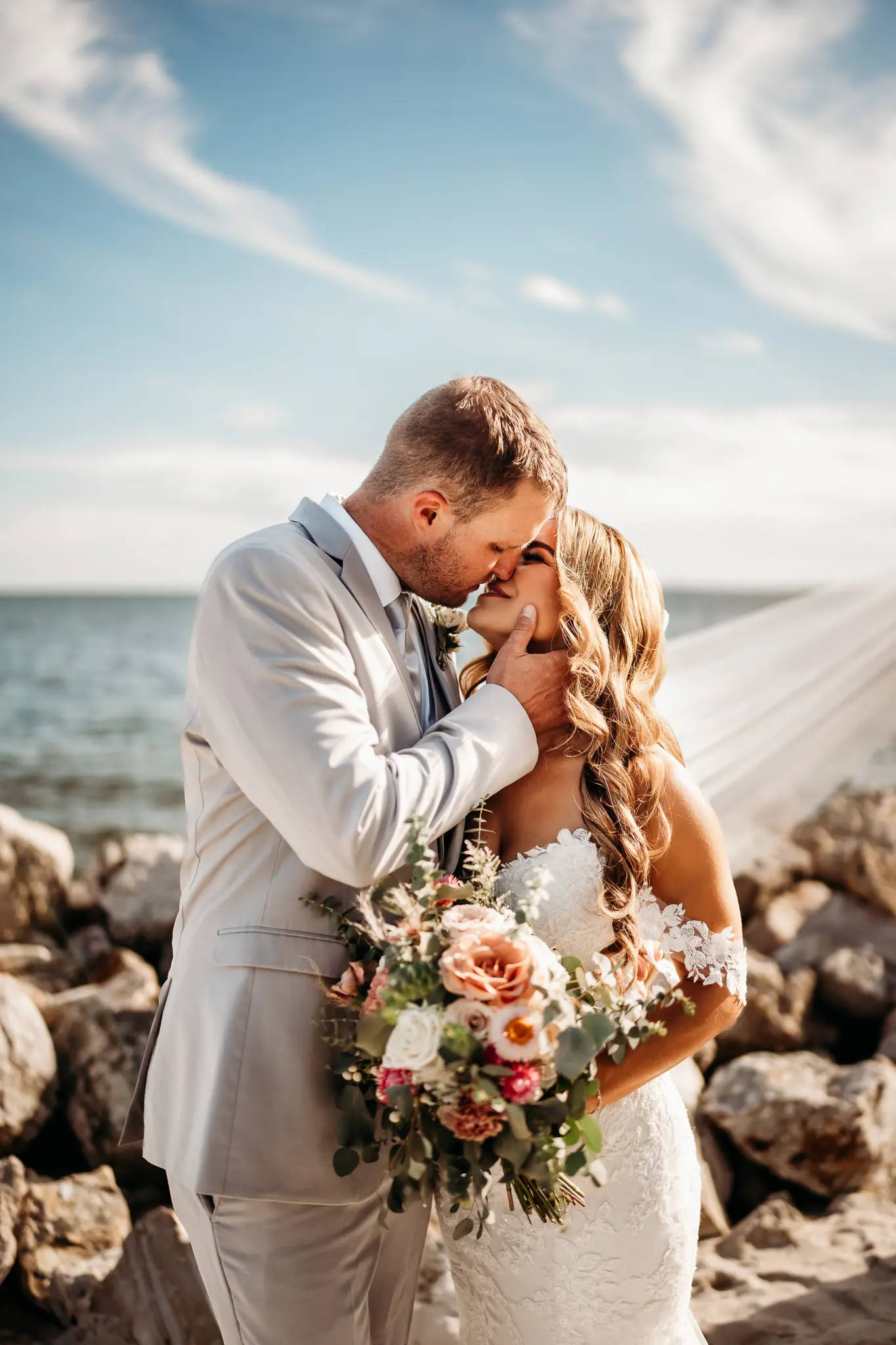 Groom and Bride Kissing on Rocks Waterfront Wedding Portrait | Fitted Casablanca Bridal Wedding Gown with Strapless Sweetheart Neckline and Floral Lace Appliques with Sequined Tulle Layer with Off the Shoulder Floral Lace Straps Inspiration | Traditional Tulle Veil Ideas | Groom Grey Suit Inspiration | Soft Beach Curls Wedding Hair Ideas | Eucalyptus and Soft White and Blush Florals Bridal Bouquet Inspiration | Tampa Bay Wedding Hair and Makeup Artist Adore Bridal Hair and Makeup | Tampa Bay Photographer Sabrina Autumn Photography