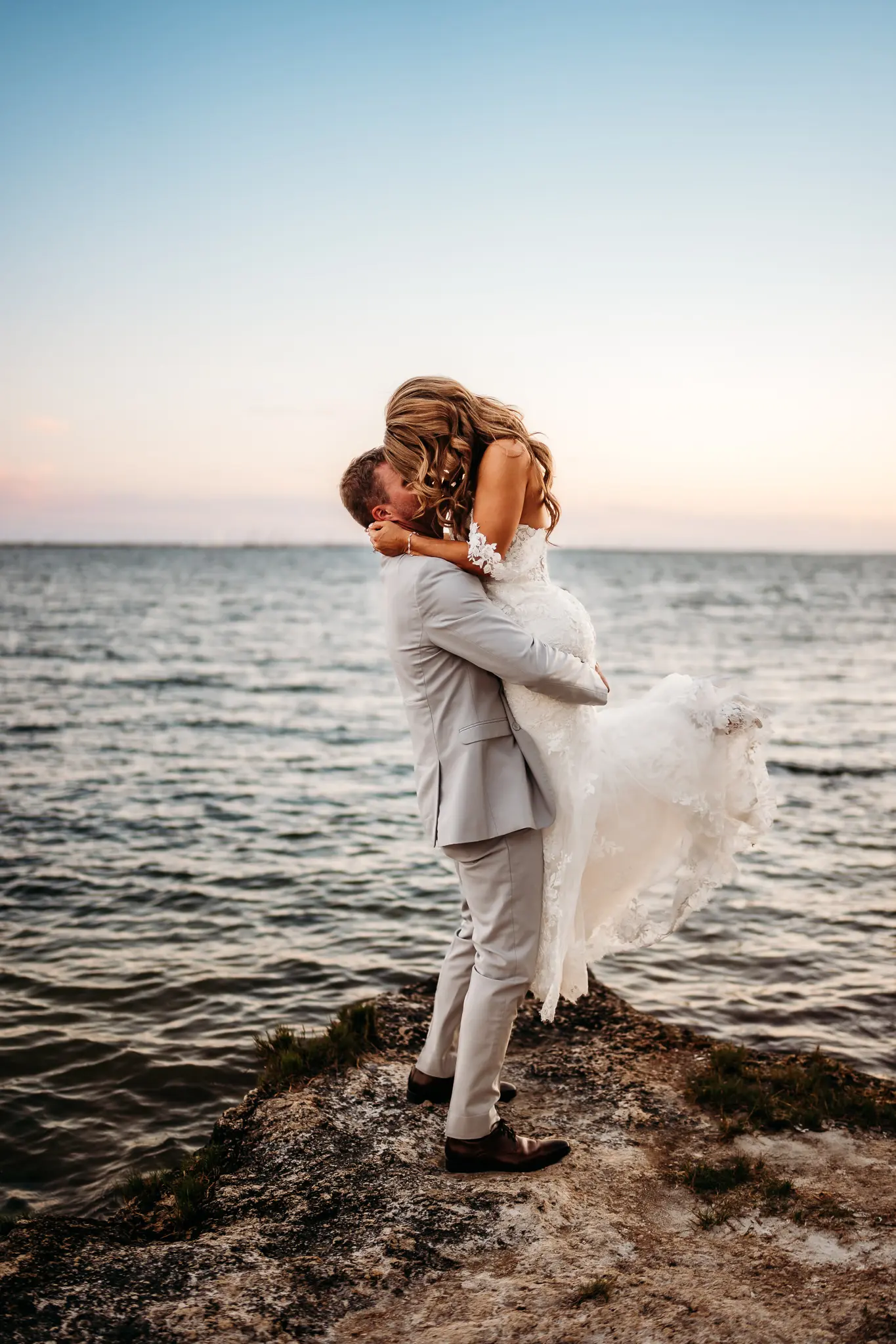 Groom Picking Up Bride Waterfront Wedding Portrait | Fitted Casablanca Bridal Wedding Gown with Strapless Sweetheart Neckline and Floral Lace Appliques with Sequined Tulle Layer with Off the Shoulder Floral Lace Straps Inspiration | Groom Grey Suit Ideas | Soft Beach Curls Wedding Hair Inspiration | Tampa Bay Wedding Hair and Makeup Artist Adore Bridal Hair and Makeup | Tampa Bay Photographer Sabrina Autumn Photography