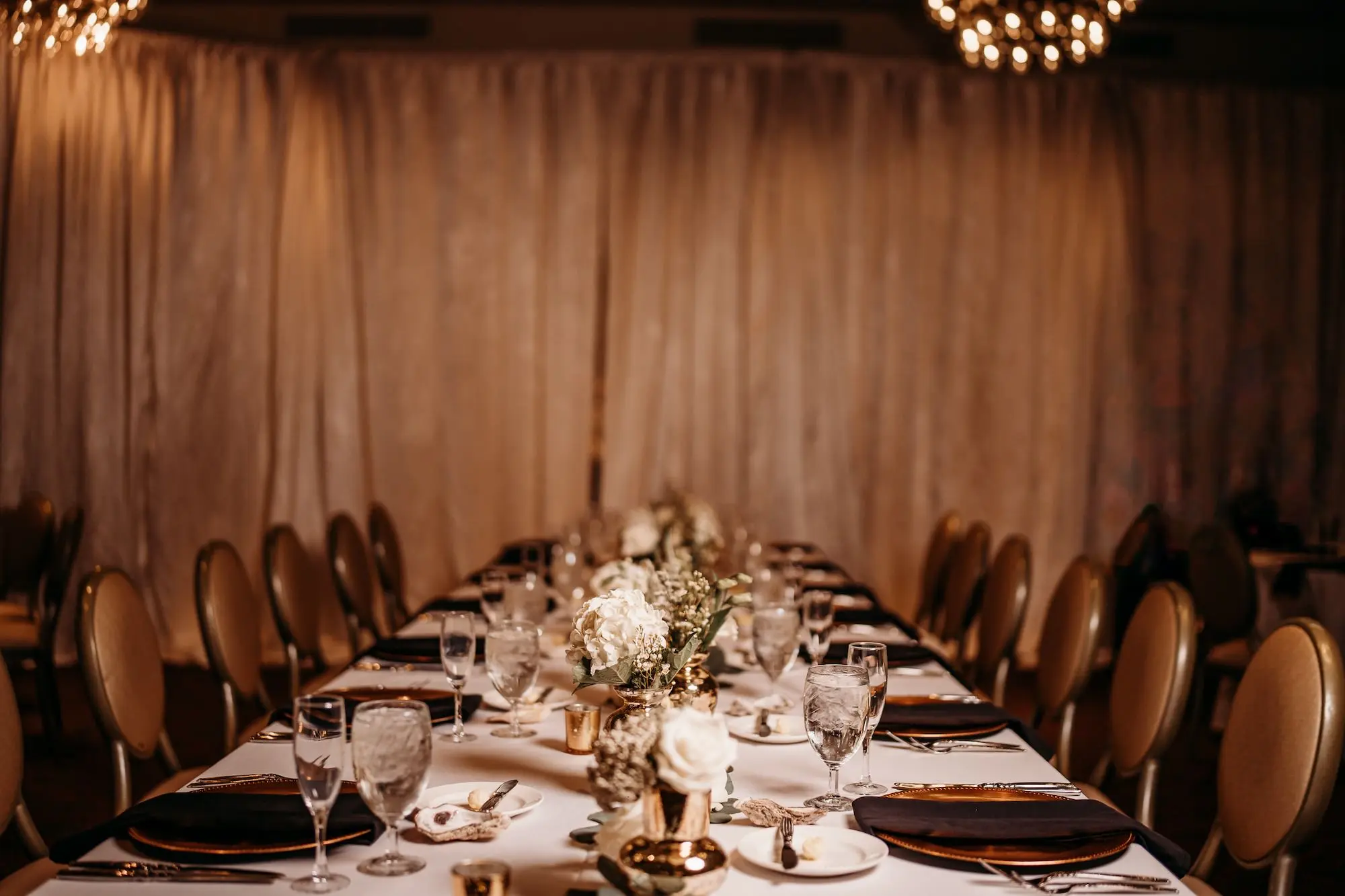 Luxurious Fall Grand Ballroom Wedding Reception Long Feasting Table Tablescape Inspiration | Gold Mercury Bud Vase Centerpieces with White Hydrangeas, Roses, and Greenery Ideas | Gold Chargers with Black Linen Napkins