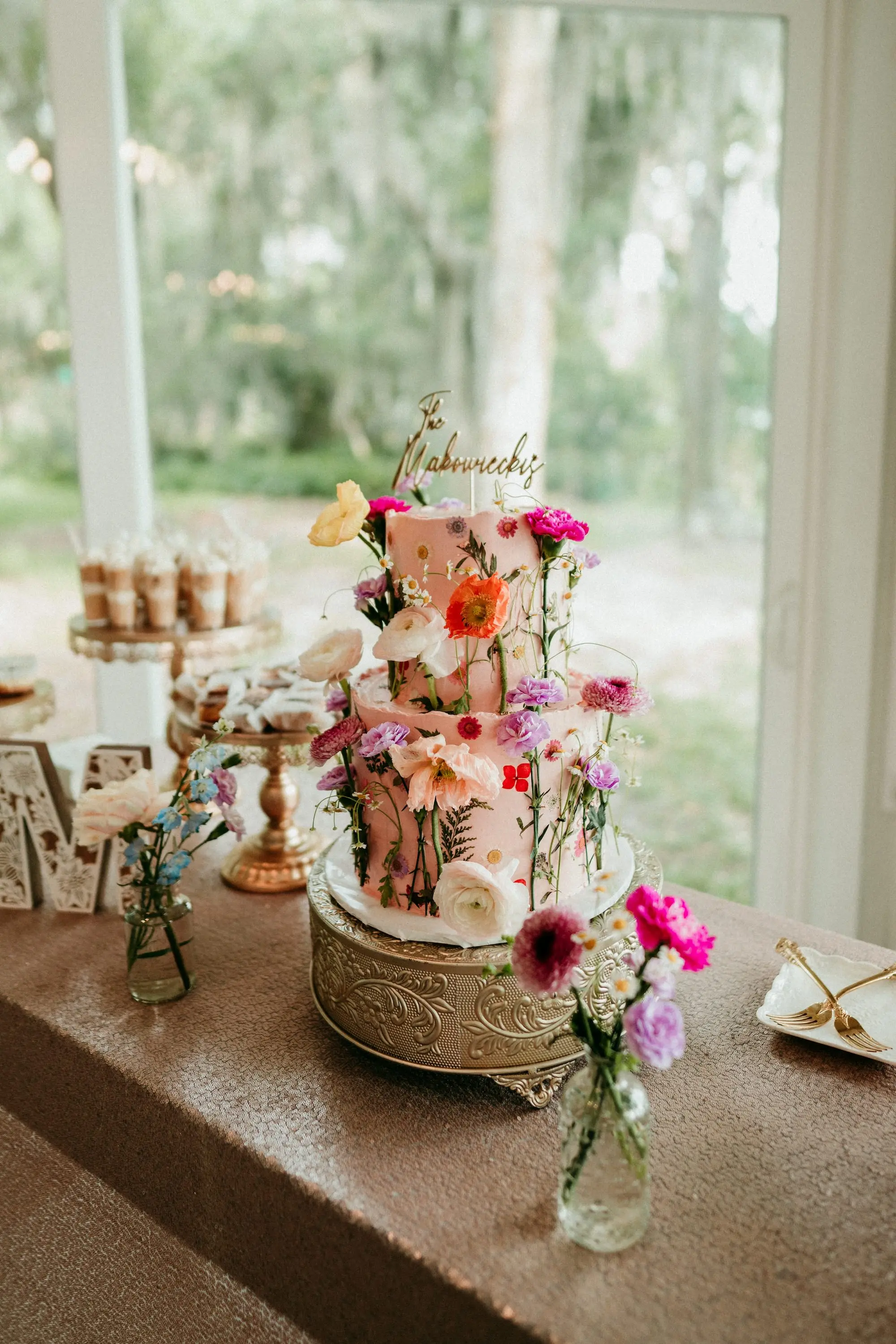 Boho Romantic Two Tier Light Pink Wedding Cake with Wildflowers on a Gold Stand and Gold Last Name Topper Inspiration | Tampa Bay Bakery Faba's Sweetshop