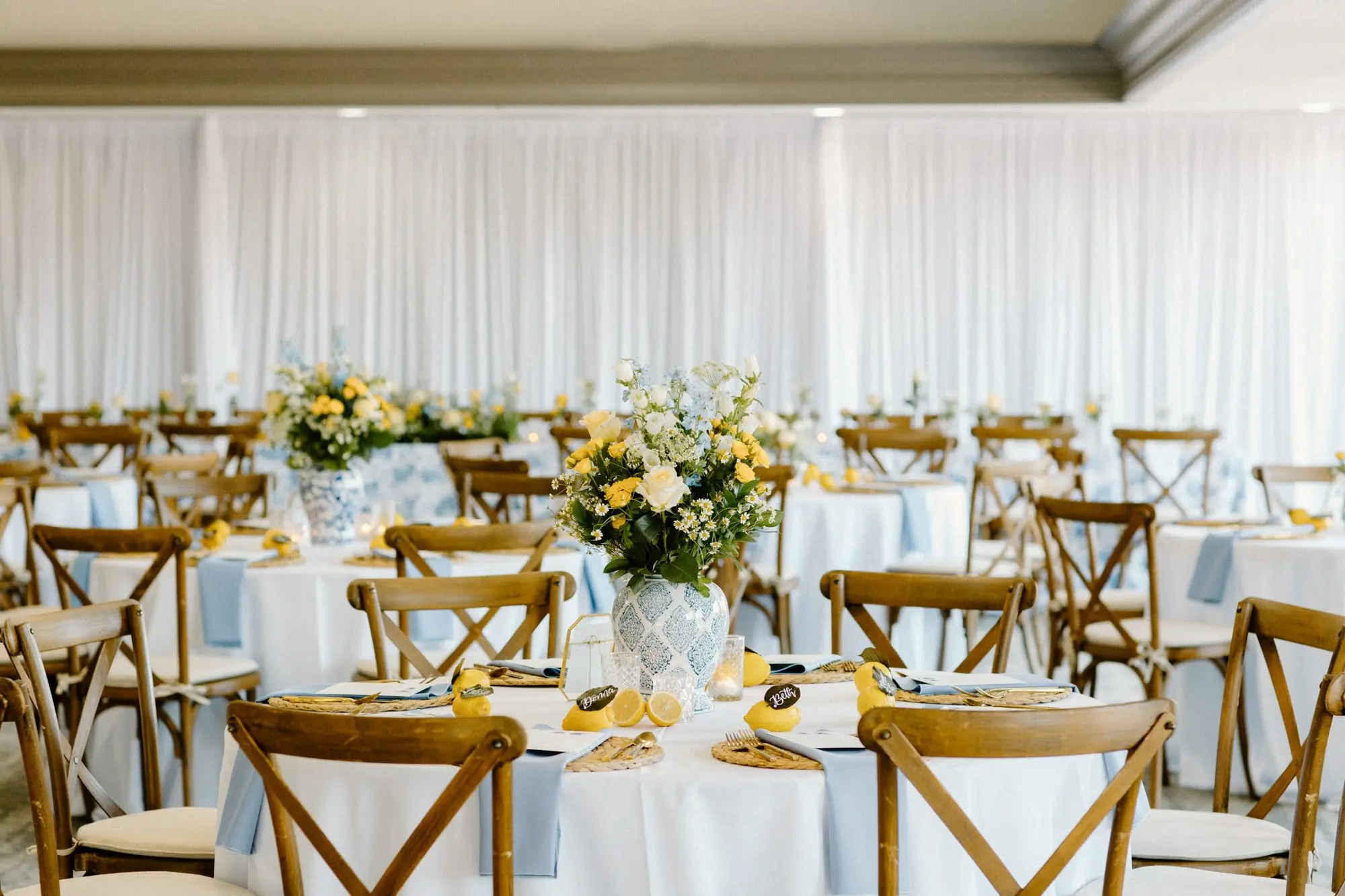 Round Tables with White Linen and Wooden Chairs Wedding Reception Decor Inspiration | Woven Chargers with Gold Flatware and Lemon Citrus Wedding Reception Decor Ideas | Blue and White Detailed Large China Vase with White Florals and Yellow Roses Centerpieces Inspiration | Tampa Bay Over The Top Linen Rentals | St Pete Rental A Chair Affair | Tampa Bay Planner Unique Weddings and Events
