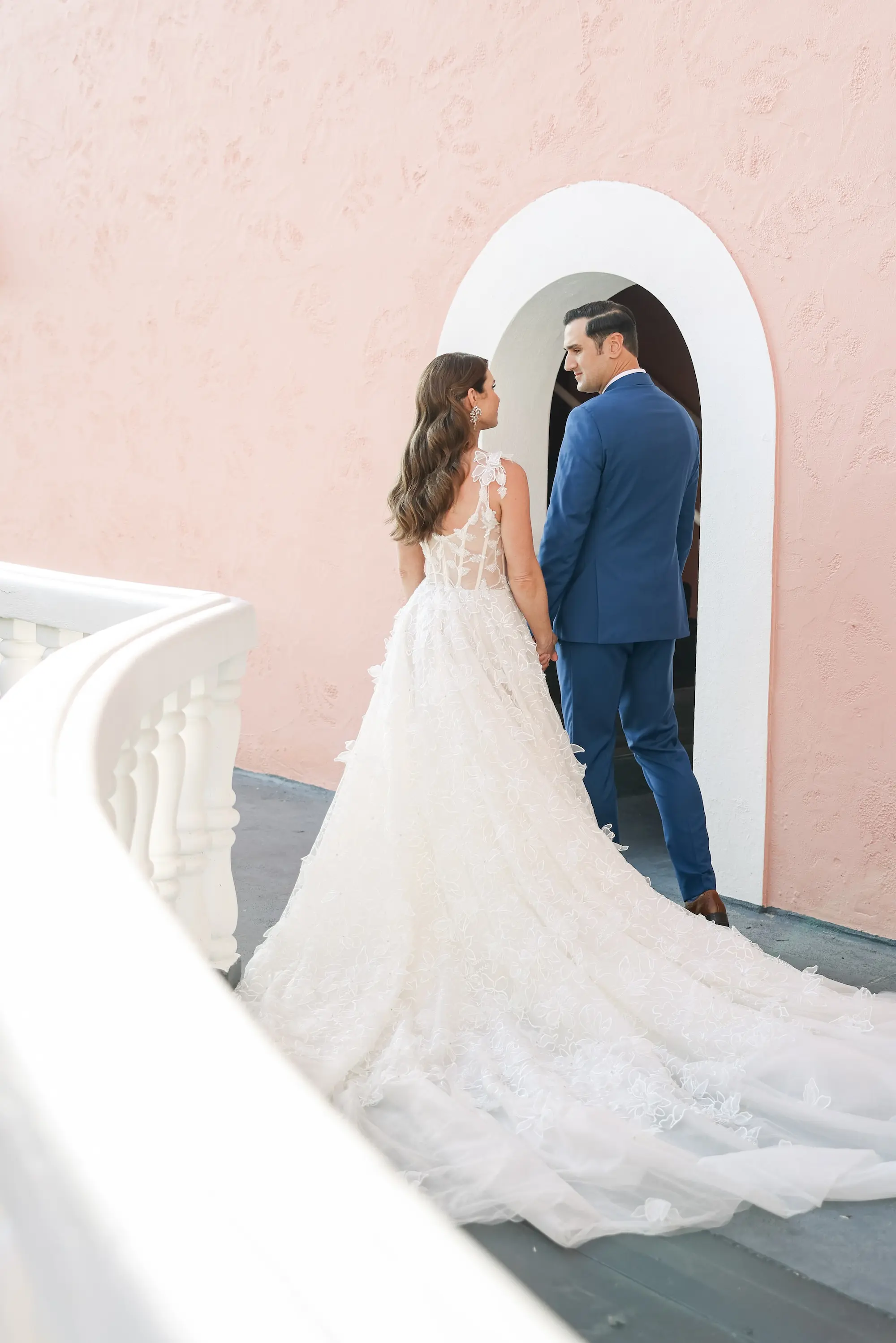 Bride and Groom Walking on Balcony Wedding Portrait | Lace and Tulle Galia Lahav Wedding Gown with Fitted Bodice and Full Layered, Ruffled Skirt and Train Ideas | Blue Groom Suit Inspiration | Soft, Romantic Beach Curls Bridal Hair Ideas | bor Hair and Makeup Artist Adore Bridal Hair and Makeup | Venue St. Pete Beach Don CeSar | Tampa Bay Planner Blue Skies Weddings and Events | St Pete Photographer Lifelong Photography Studio