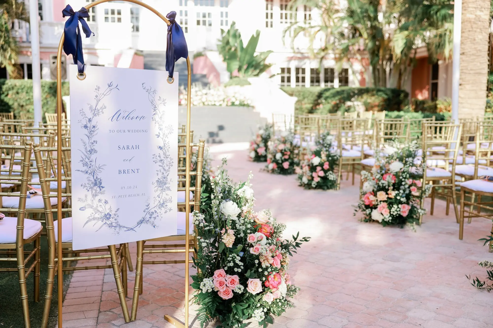 White Cardstock with Blue Floral Details Welcome Wedding Ceremony Sign on Gold Arched Frame with Navy Satin Ribbons Decor Inspiration | Pink, White, Coral Roses and Baby's Breath Flower Arrangement Ceremony Aisle Decor Ideas | Gold Chiavari Chair Rentals by Tampa Bay Kate Ryan Event Rentals | Outdoor Beachfront Resort Wedding Ceremony Venue | St Pete Beach Wedding Venue Don CeSar | Tampa Bay Planner Blue Skies Weddings and Events