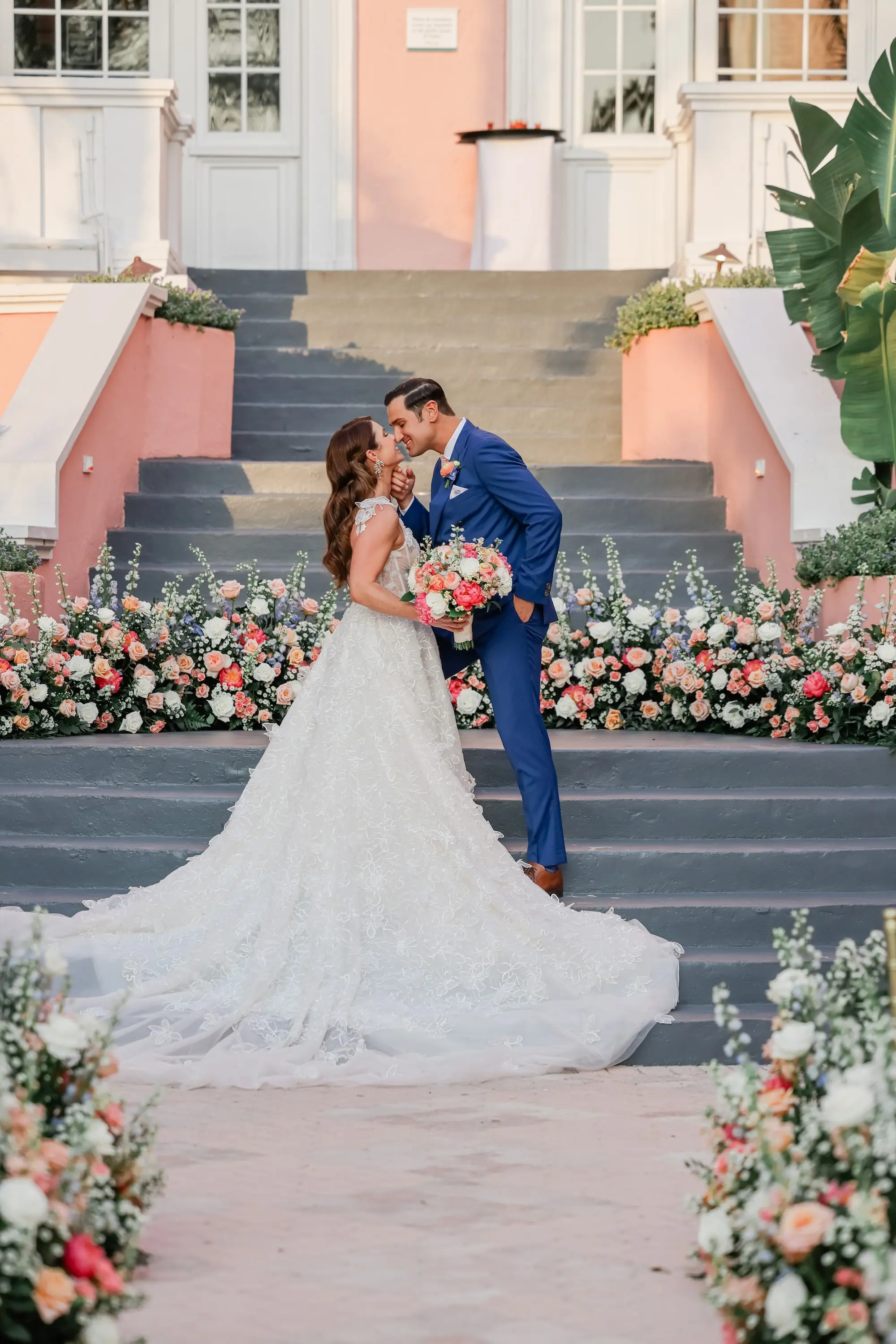 Bride and Groom on Steps of Historic Pink Palace St Pete Beach Wedding Venue Don CeSar | White, Pink, Coral, Peach, Blush Roses, Peonies, Carnation and Wisteria Flower Arrangement Wedding Ceremony Decor Inspiration | Lace and Tulle Galia Lahav Wedding Gown with Fitted Bodice and Full Layered, Ruffled Skirt and Train Ideas | Soft, Curls Bridal Hair Inspiration | Soft Beach Curls Bridal Hair Ideas | Ybor Hair and Makeup Artist Adore Bridal Hair and Makeup | St Pete Beach Resort Wedding Venue | Tampa Bay Planner Blue Skies Weddings and Events | St Pete Photographer Lifelong Photography Studio