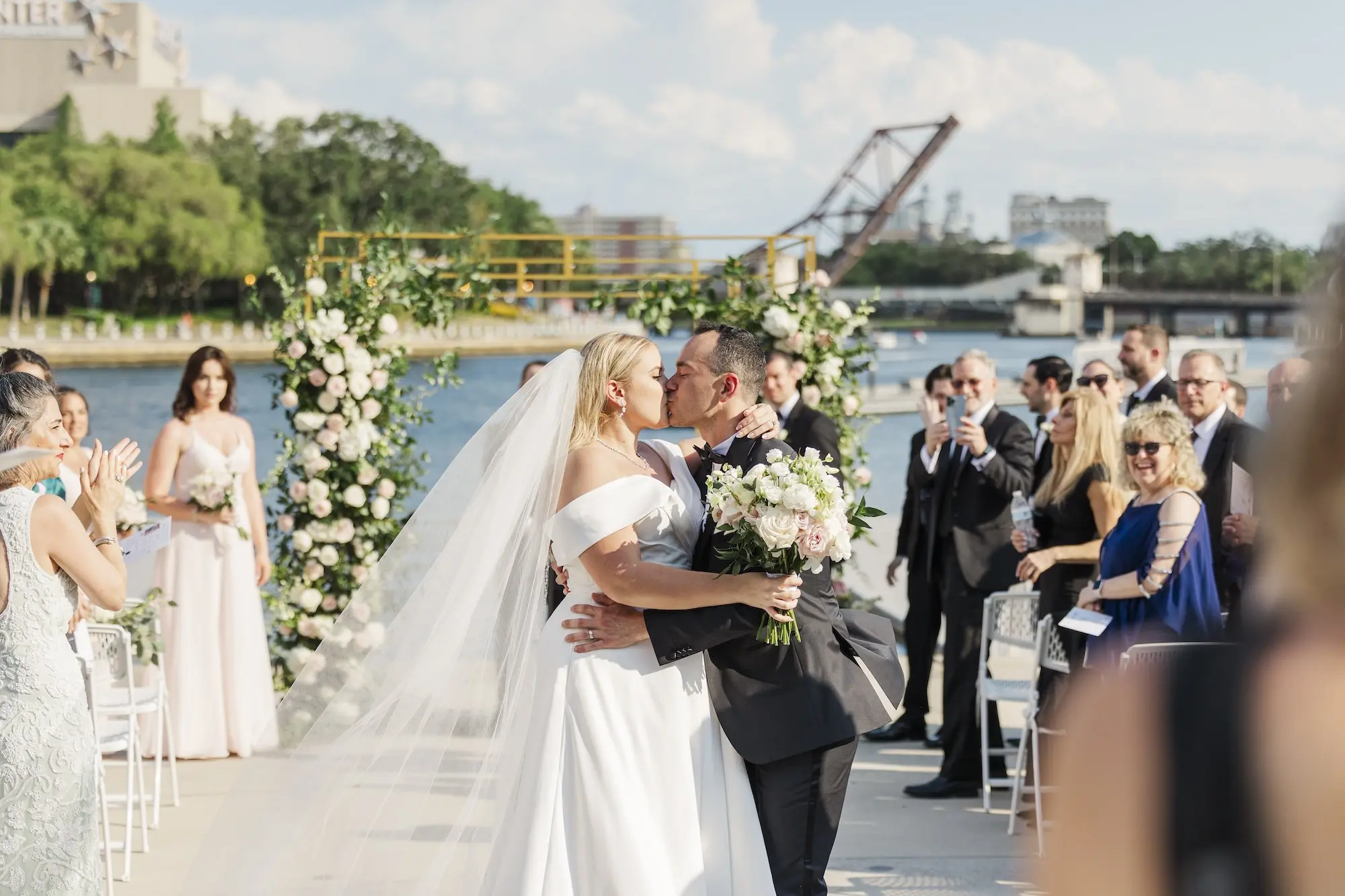 Bride and Groom Kiss in Wedding Ceremony Aisle Portrait | Romantic Outdoor Riverfront Wedding Ceremony Inspiration | Gold Chuppah with Light Pink and White Cascading Florals and Greenery Ideas | Tampa Bay Florist Save The Date Florida | Florida Photographer Mary Anna Photography | Venue Tampa River Center
