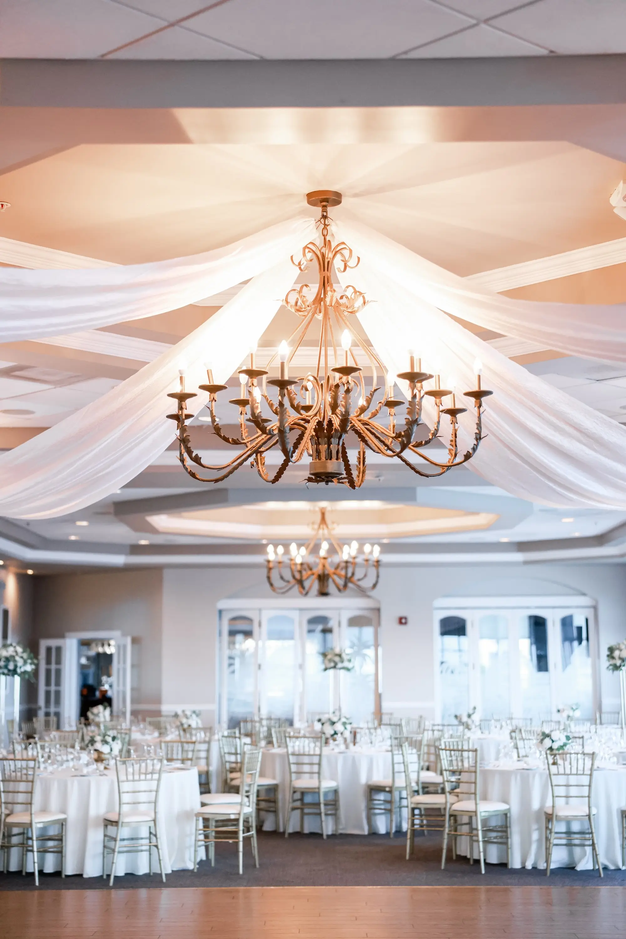 Luxurious Ballroom Wedding Reception | Gold Candelabra Chandelier with White Hanging Ceiling Draping | Round Wedding Reception Tables with White Linens and Chiavari Chairs | Short Classic Centerpieces with White Roses and Greenery Flower Arrangement Ideas | St Pete Event Venue Isla Del Sol Yacht and Country Club | Tampa Bay Planner Planner Coastal Coordinating
