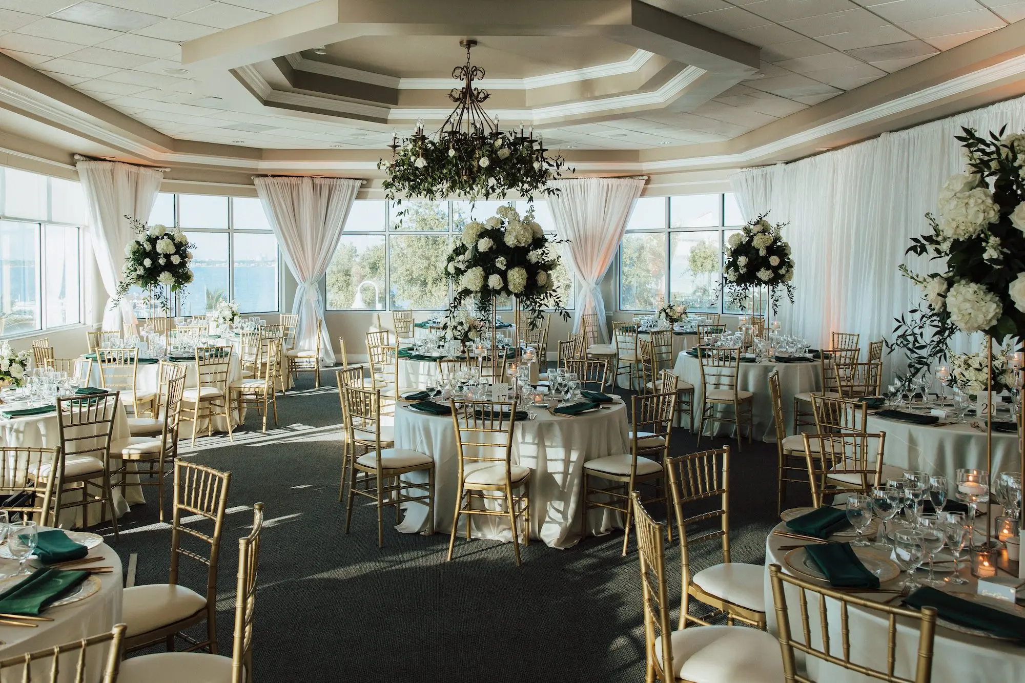 Luxurious Waterfront Ballroom Wedding Reception | Gold Chiavari Wedding Chairs at Round Table with White Linens and Dark Green Napkins Decor Ideas | Tall Gold Stands with Lush White and Cream with Greenery Floral Wedding Reception Table Centerpieces Inspiration | Tampa Bay Kate Ryan Event Rentals | Tampa Bay Event Venue Isla Del Sol Yacht and Country Club