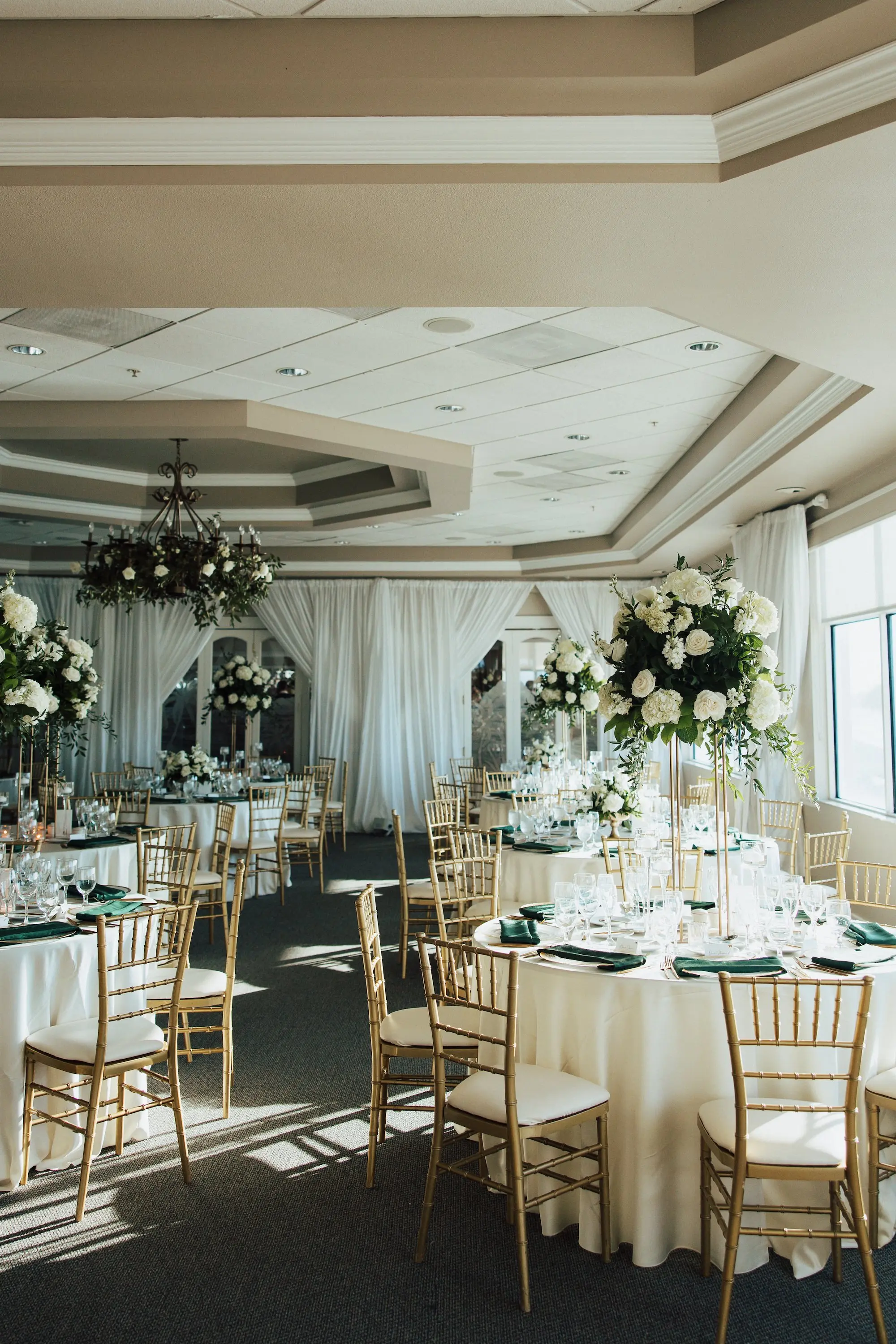 Luxurious Classic White Garden Theme Country Club Ballroom Wedding Reception | Gold Chiavari Wedding Chairs at Round Table with White Linens and Dark Green Napkins Decor Ideas | Tall Gold Stands with Lush White and Cream with Greenery Floral Wedding Reception Table Centerpieces Inspiration | Tampa Bay Kate Ryan Event Rentals | St Pete Event Venue Isla Del Sol Yacht and Country Club