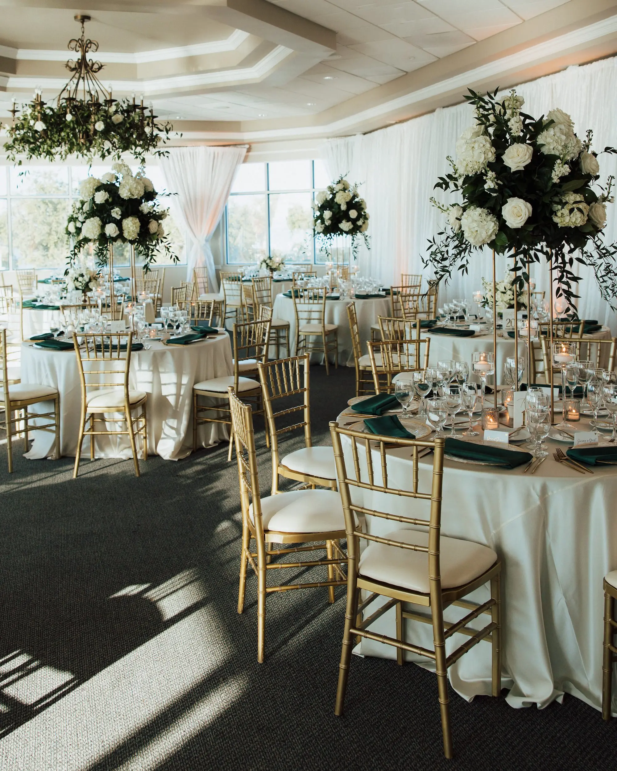Luxurious Waterfront Ballroom Wedding Reception | Gold Chiavari Wedding Chairs at Round Table with White Linens and Dark Green Napkins Decor Ideas | Tall Gold Stands with Lush White and Cream with Greenery Floral Wedding Reception Table Centerpieces Inspiration | Tampa Bay Kate Ryan Event Rentals | Tampa Bay Event Venue Isla Del Sol Yacht and Country Club