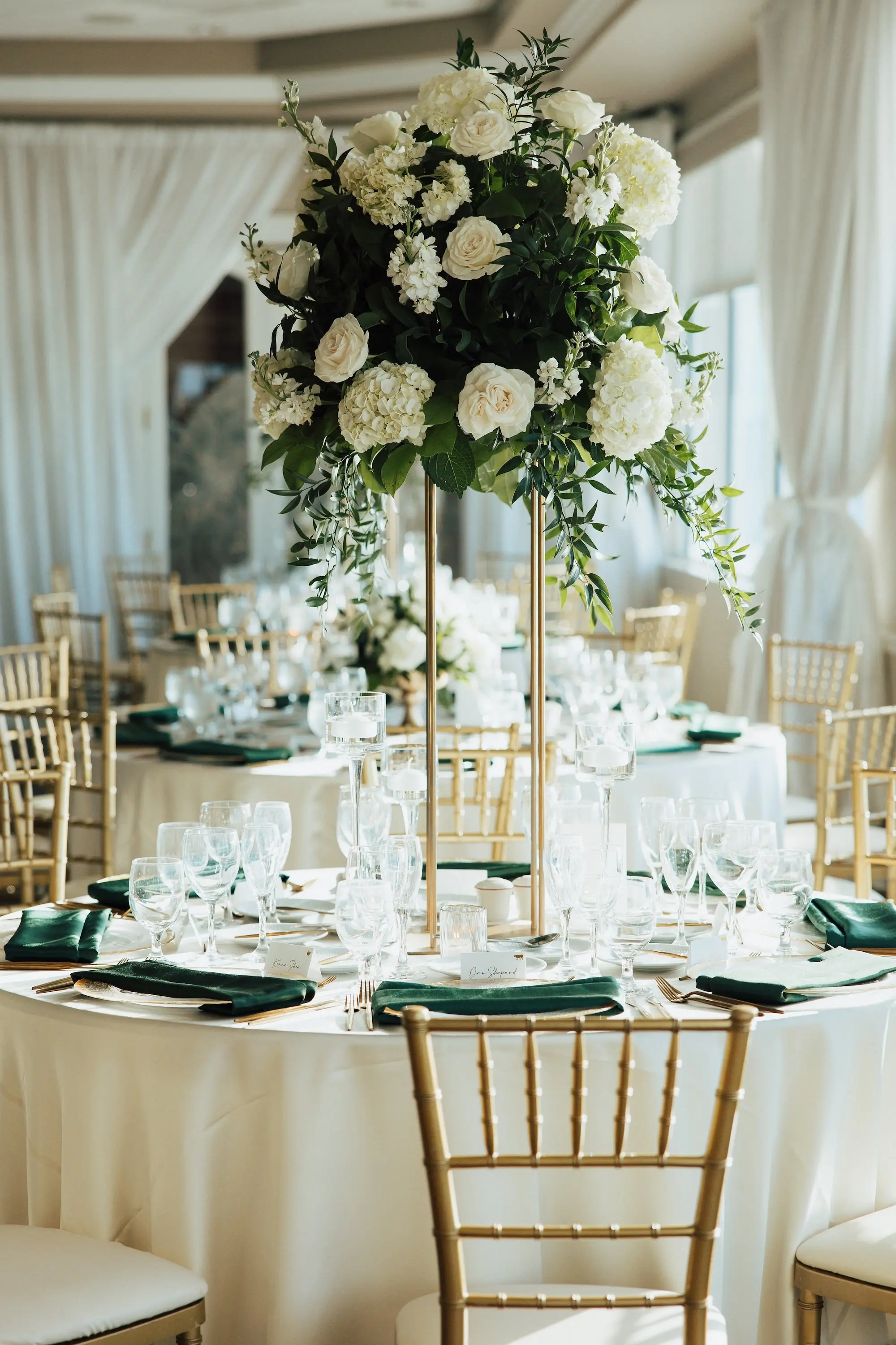 Luxurious Waterfront Ballroom Wedding Reception | Gold Chiavari Wedding Chairs at Round Table with White Linens and Dark Green Napkins Decor Ideas | Tall Gold Stands with Lush White and Cream with Greenery Floral Wedding Reception Table Centerpieces Inspiration | Tampa Bay Kate Ryan Event Rentals | Tampa Bay Event Venue Isla Del Sol Yacht and Country Club