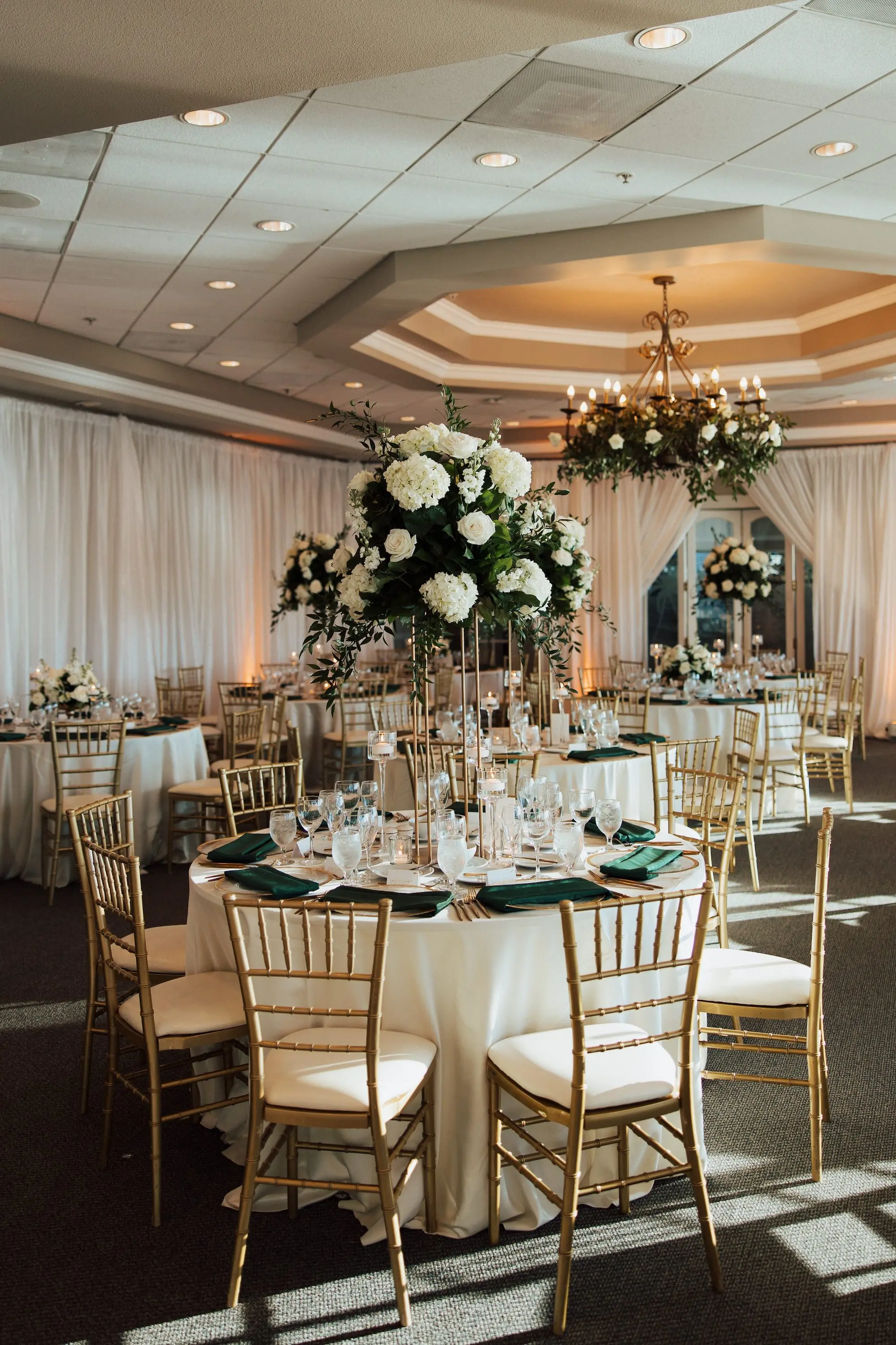 Luxurious Classic White Garden Theme Country Club Ballroom Wedding Reception | Gold Chiavari Wedding Chairs at Round Table with White Linens and Dark Green Napkins Decor Ideas | Tall Gold Stands with Lush White and Cream with Greenery Floral Wedding Reception Table Centerpieces Inspiration | Tampa Bay Kate Ryan Event Rentals | Tampa Bay Event Venue Isla Del Sol Yacht and Country Club