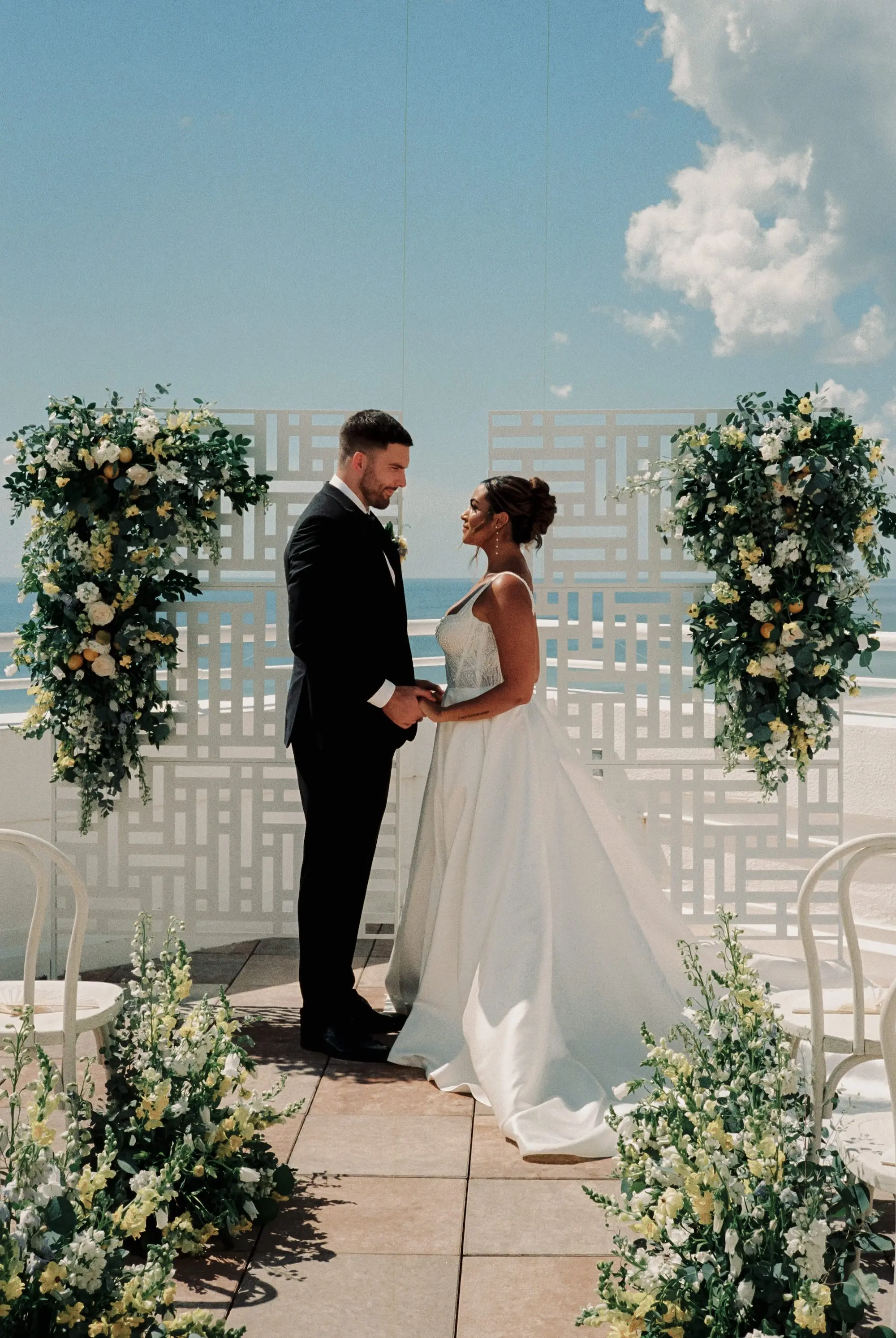 Bride and Groom Wedding Ceremony Vow Exchange Wedding | Outdoor Beachfront Wedding Ceremony Venue Inspiration | White and Yellow Roses and Florals in Lush Greenery Altar Ceremony Decor Ideas | Classic Groom Black and White Tuxedo Inspiration | Satin Ballgown with Square Neckline and Slit Wedding Dress Boutique Truly Forever Bridal Tampa | Photographer Amber Yonker Photography | St Pete Beach Wedding Venue Don CeSar