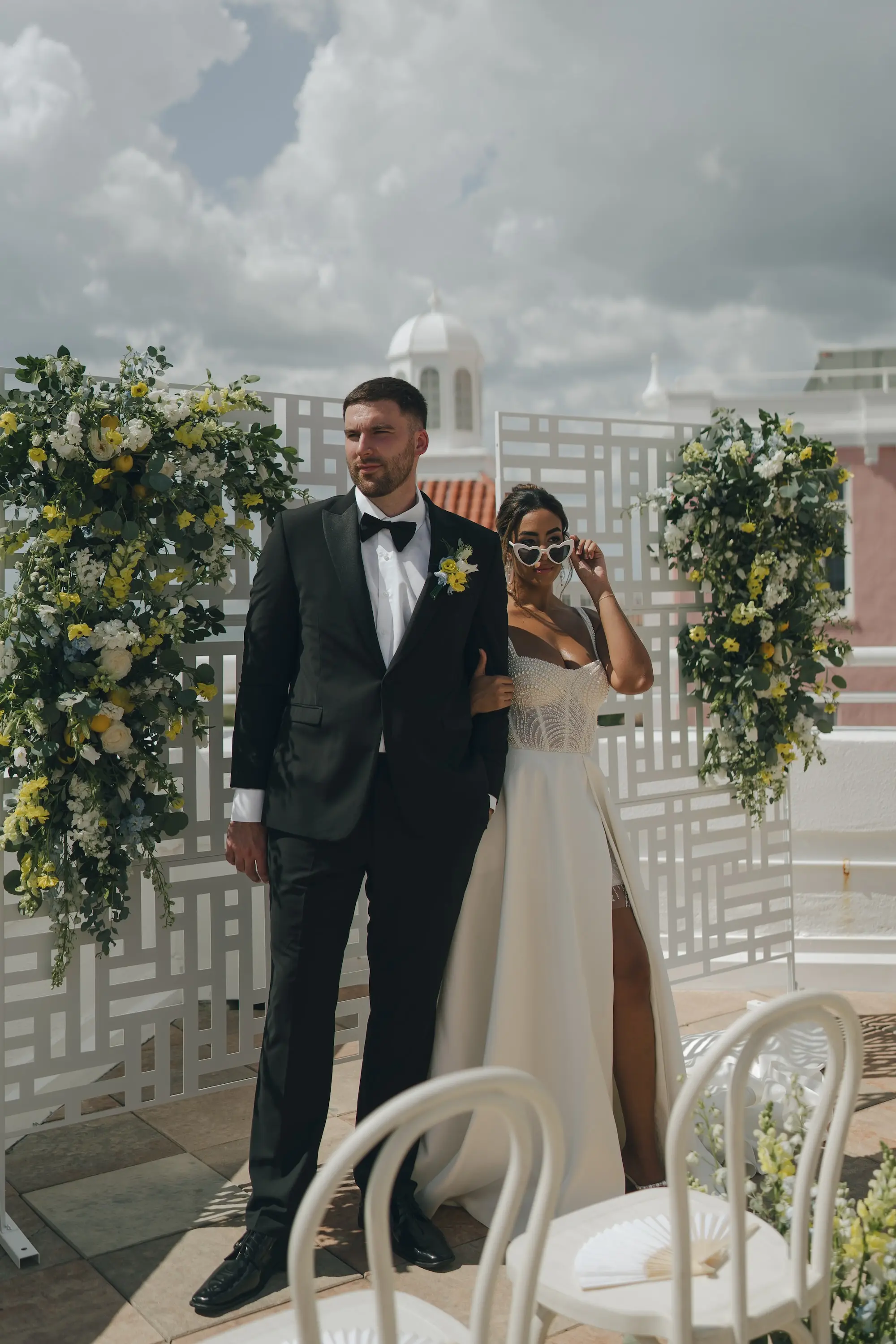 Bride and Groom Balcony Wedding Portrait | Outdoor Beachfront Wedding Ceremony Venue Inspiration | White and Yellow Roses and Florals in Lush Greenery Altar Ceremony Decor Ideas | Classic Groom Black and White Tuxedo Inspiration | Satin Ballgown with Corset Top and Slit Wedding Dress Boutique Truly Forever Bridal Tampa | Updo with Loose Beach Curls Wedding Hair Inspiration | Romantic Bridal Wedding Hair Updo and Makeup Ideas | Hair and Makeup Artist Lasting Luxe | Photographer Amber Yonker Photography | St Pete Beach Wedding Venue Don CeSar