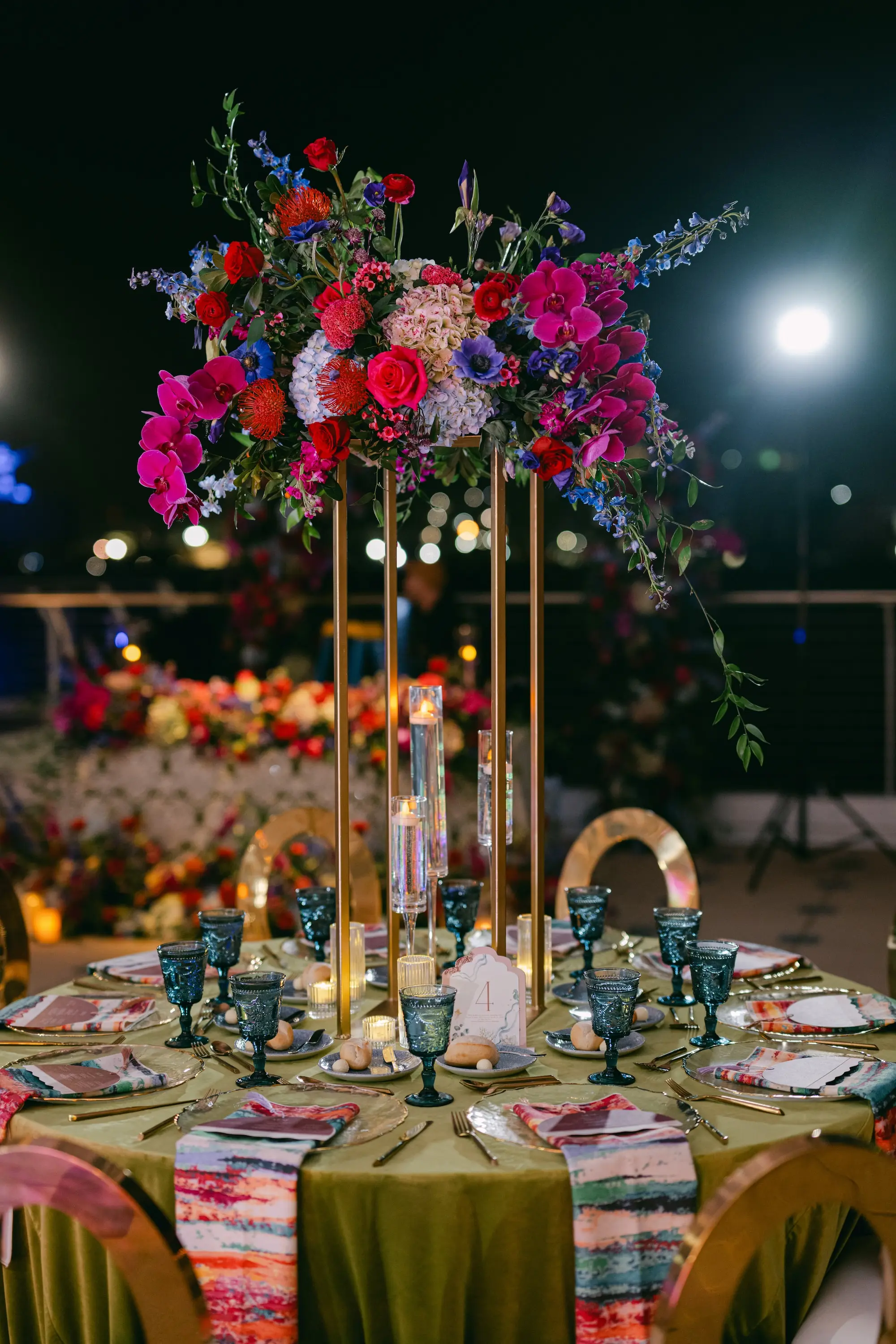 Vibrant Jewel-Toned Wedding Reception Tablescape Decor Ideas | Pink, Blue, and Purple Roses, Orchids, Hydrangeas, Wisteria Floral Arrangement on Gold Centerpiece Inspiration | Olive Green Linen Tablecloth with Pattern Cloth Napkin Wedding Reception Decor Ideas | Blue Depression Glass Drinkware and Silver Flatware Inspiration | Different Size Vases for Floating Tea Candles Reception Decor Ideas | Tampa Bay Planner Blue Skies Weddings and Events