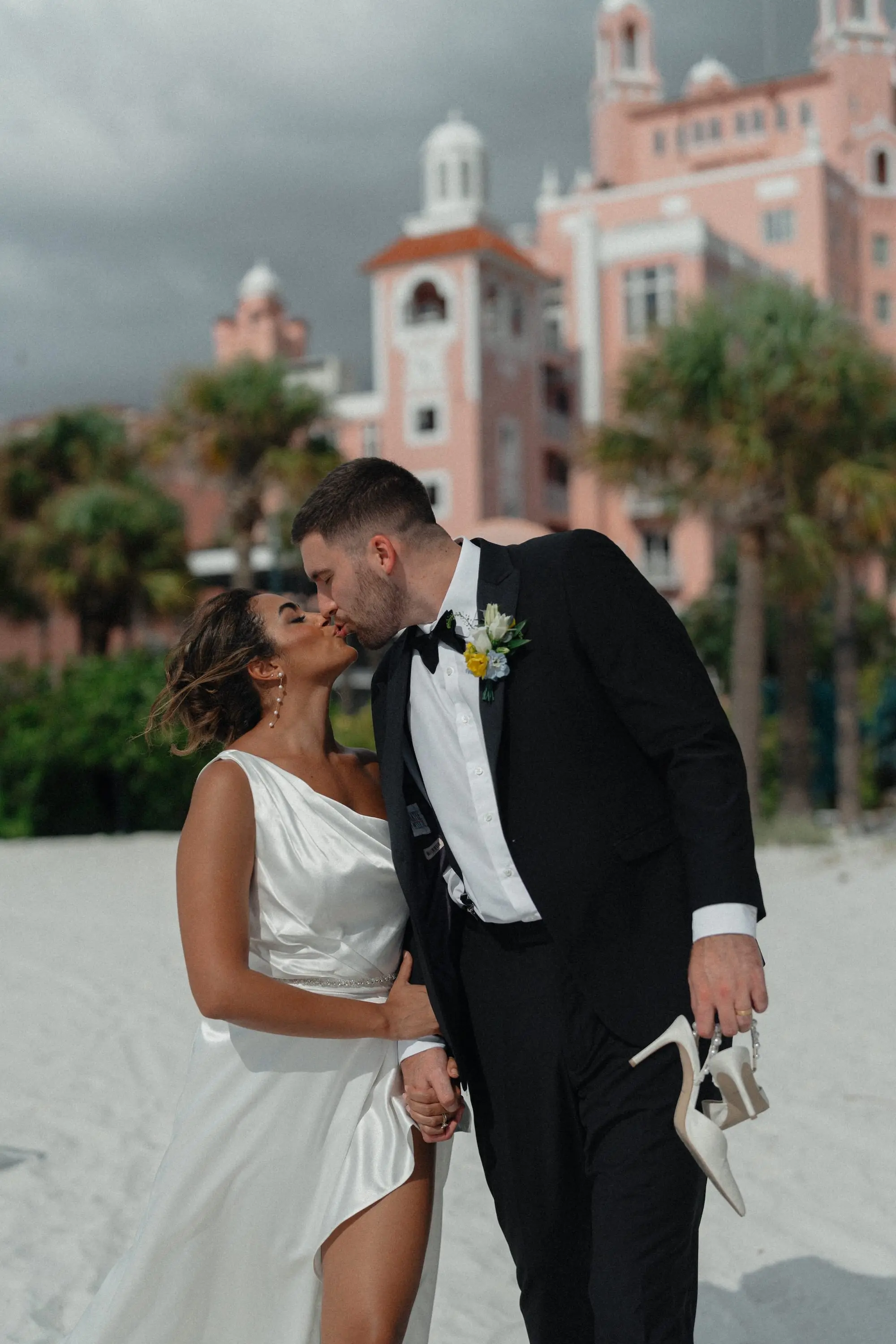 Bride and Groom Kissing on the Beach with Groom Holding Bride Shoes Wedding Portrait | Satin One Shoulder Wedding Gown Inspiration | Photographer Amber Yonker Photography | Boutique Truly Forever Bridal Tampa | Historic St Pete Beach Wedding Venue Don CeSar