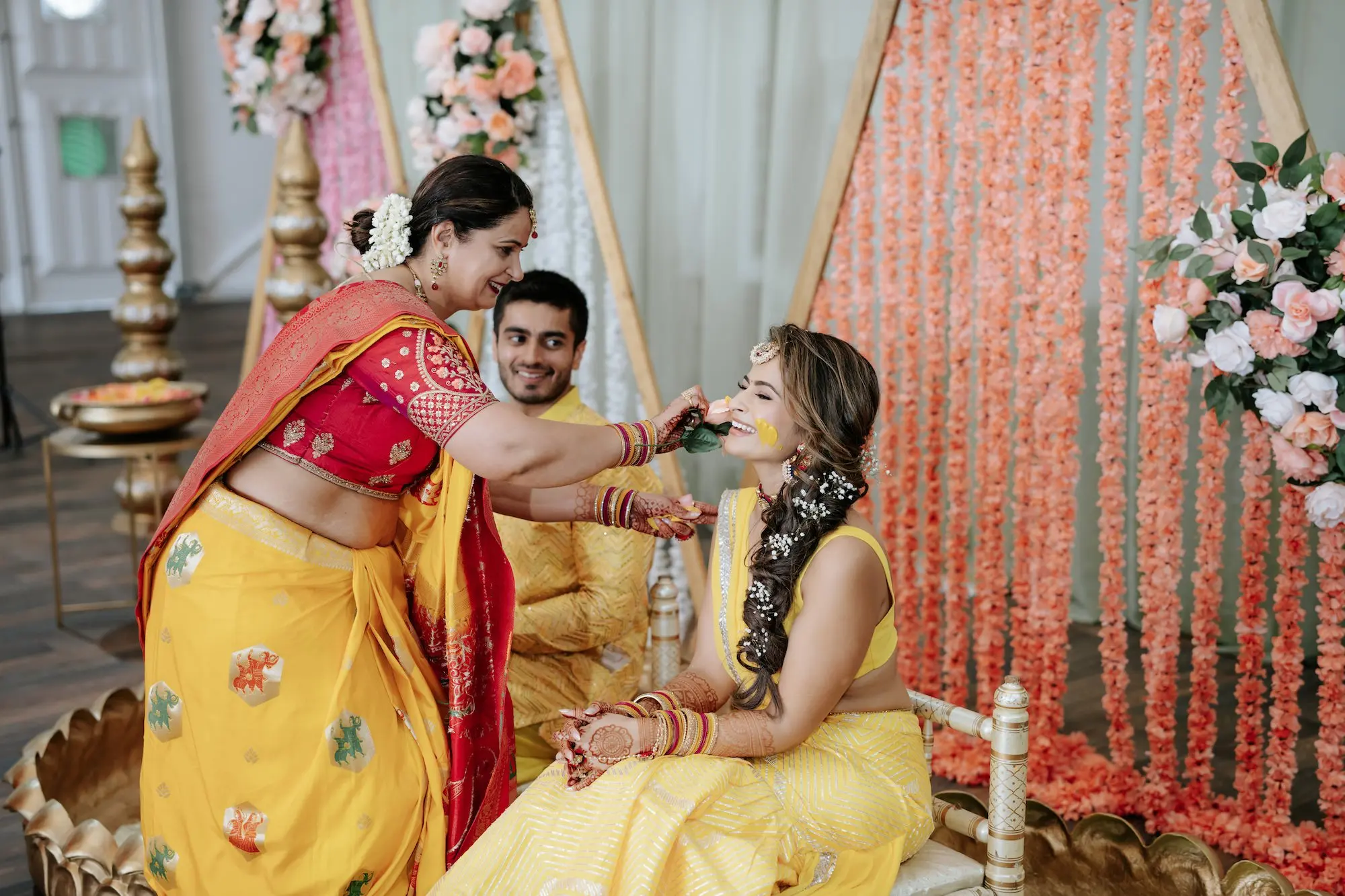Bride and Groom Haldi Wedding Ceremony Inspiration | Traditional Indian Wedding Clothing Yellow Lehenga Choli and Kurta Pajama Set Ideas | Haldi Ceremony Turmeric Paste Inspiration | Orange, Pink, and White Marigolds Hanging from Triangle Shape Backdrop Ideas | White, Blush and Pink Rose Flower Arrangement Wedding Decor Inspiration