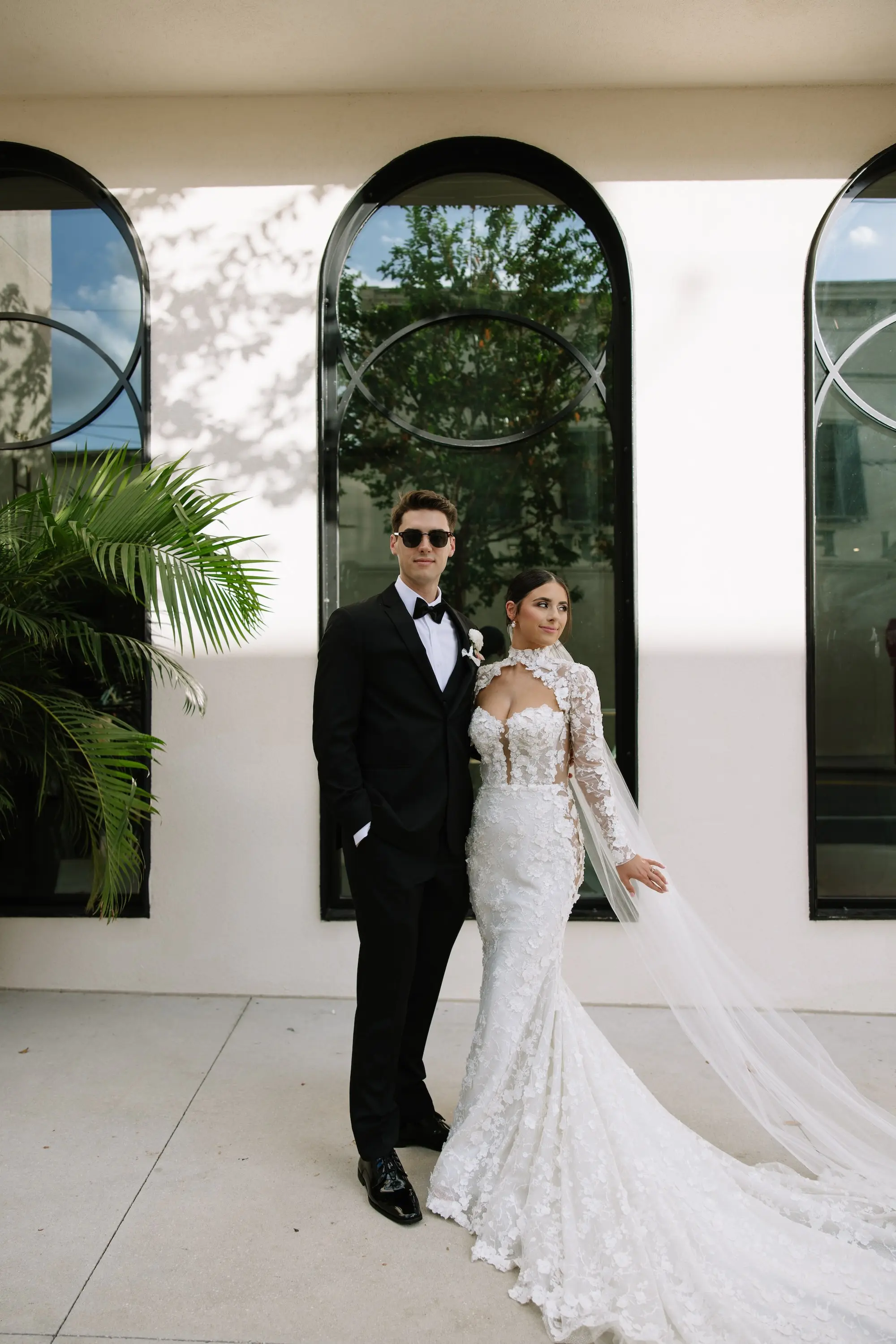 Bride and Groom Outside Hotel Haya Wedding Portrait | Intricate Floral Lace Fitted Silhouette Liz Young Wedding Gown with Long Sleeves, High Neckline with a Bolero-Style Lace Overlay and Long Train Inspiration | Traditional Chapel-Length Tulle Veil Ideas | Classic Black and White Groom Tuxedo Inspiration