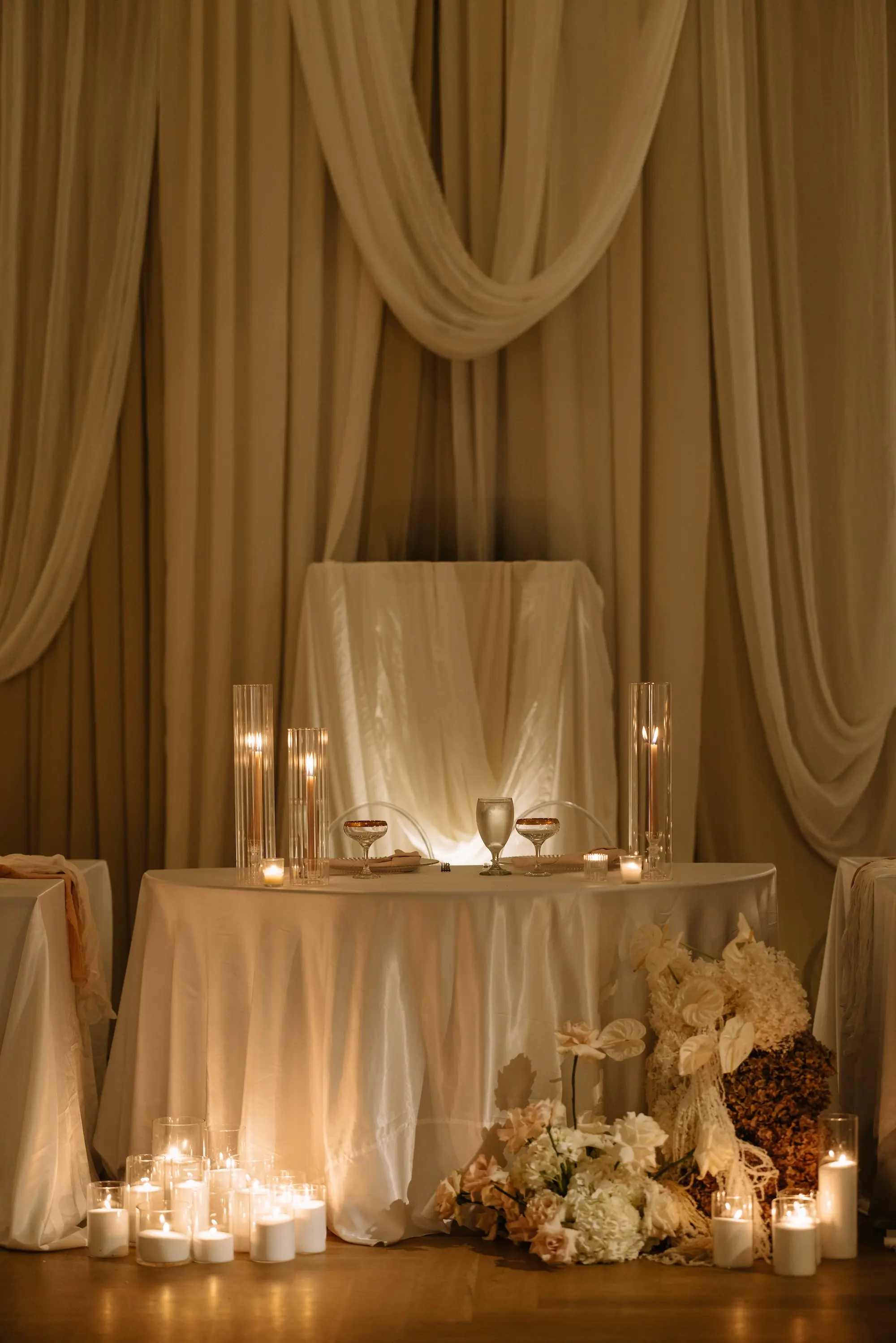 Luxurious Ballroom Wedding Reception Sweetheart Table Inspiration | White Linen Tablecloth with Romantic Taper Candles and Champagne Coupes Ideas | Ethereal Wall Draping Fabric and Large Candles in Clear Vases Decor Inspiration | Neutral Flower Arrangement Decor with White, Blush, Brown Hydrangeas, Carnations and Roses Ideas | Tampa Bay Planner EventFull Weddings