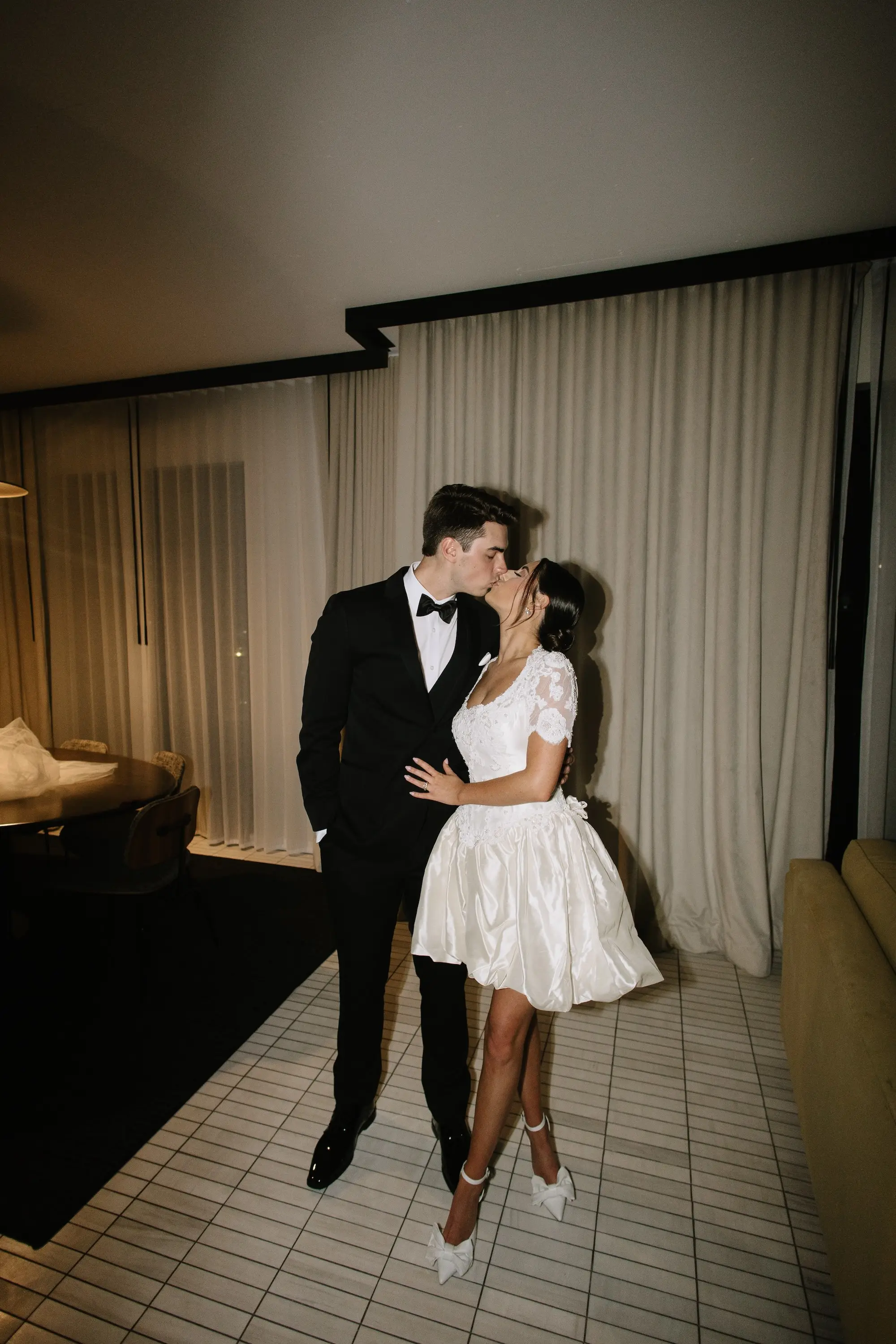Bride and Groom Kissing Wedding Reception Portrait | Short White A-line Reception Dress with Fitted Bodice, Lace Sleeves and Flared Skirt Inspiration | White Ankle Strap High Heels with Large Bow Ideas