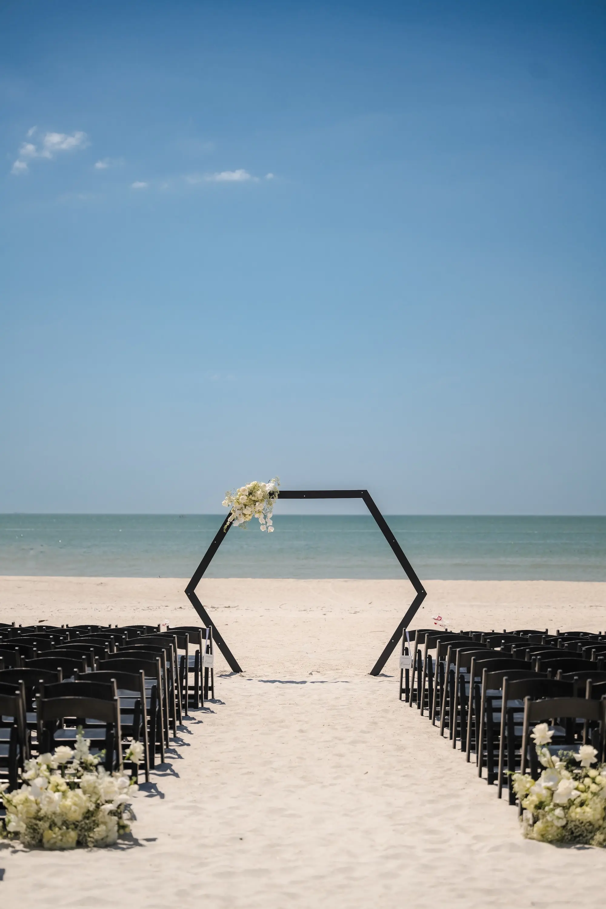 Classic Champagne and Black Florida Beachfront Wedding Ceremony Decor Inspiration | Black Wooden Ceremony Chairs Ideas | White Roses and Baby's Breath Flower Arrangement Ceremony Aisle Decor Ideas | Beach Wedding Decor Inspiration | Black Hexagon Altar Backdrop Decor Ideas | Tampa Bay Event Venue Tradewinds Island Resort
