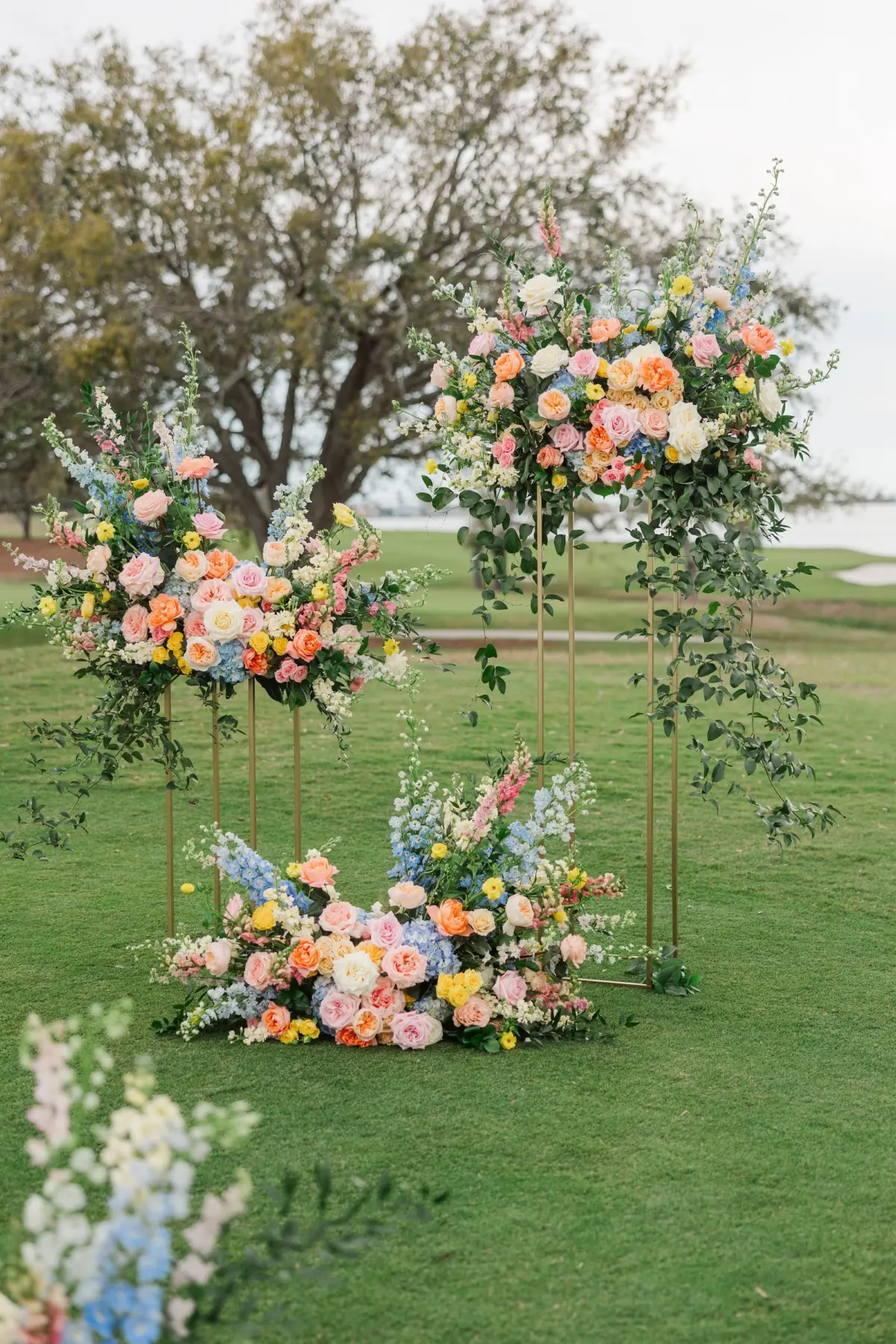 Bridgerton Spring Outdoor Garden Party Wedding Ceremony Decor Ideas | Pastel Blue, Pink, White, Yellow, and Orange Flower Arrangement on Gold Stands Altar Decor Inspiration | Country Club Golf Course Lawn Ceremony Ideas | Clearwater Florist Bruce Wayne Florals
