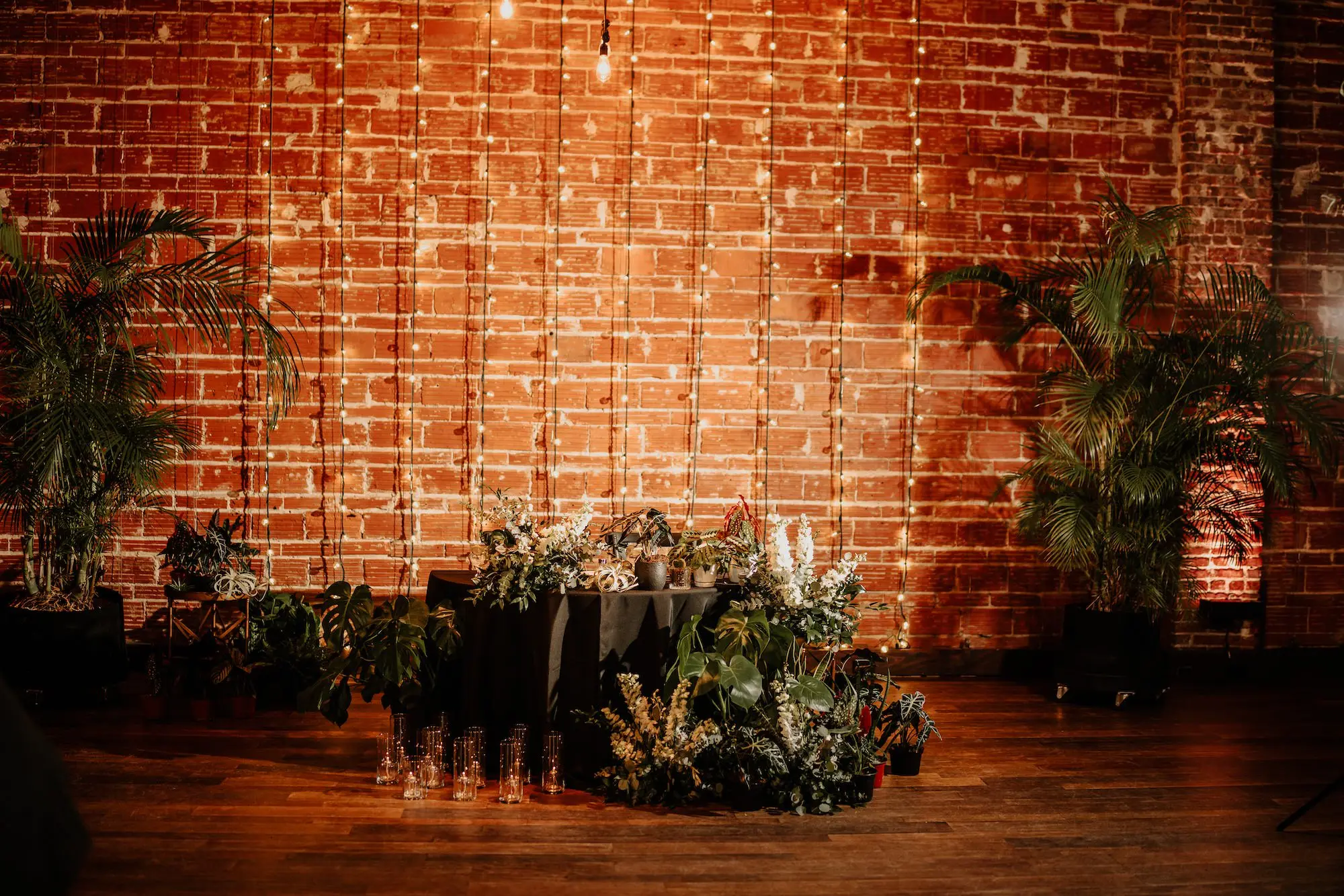 Moody, Romantic Wedding Reception Sweetheart Table Wedding Decor with Green Palms and White Florals Inspiration | String Light Backdrop on Exposed Brick Wall Ideas | Plant Wedding Themed Inspiration | Tampa Bay Premier Wedding Venue NOVA 535 in Downtown St. Pete