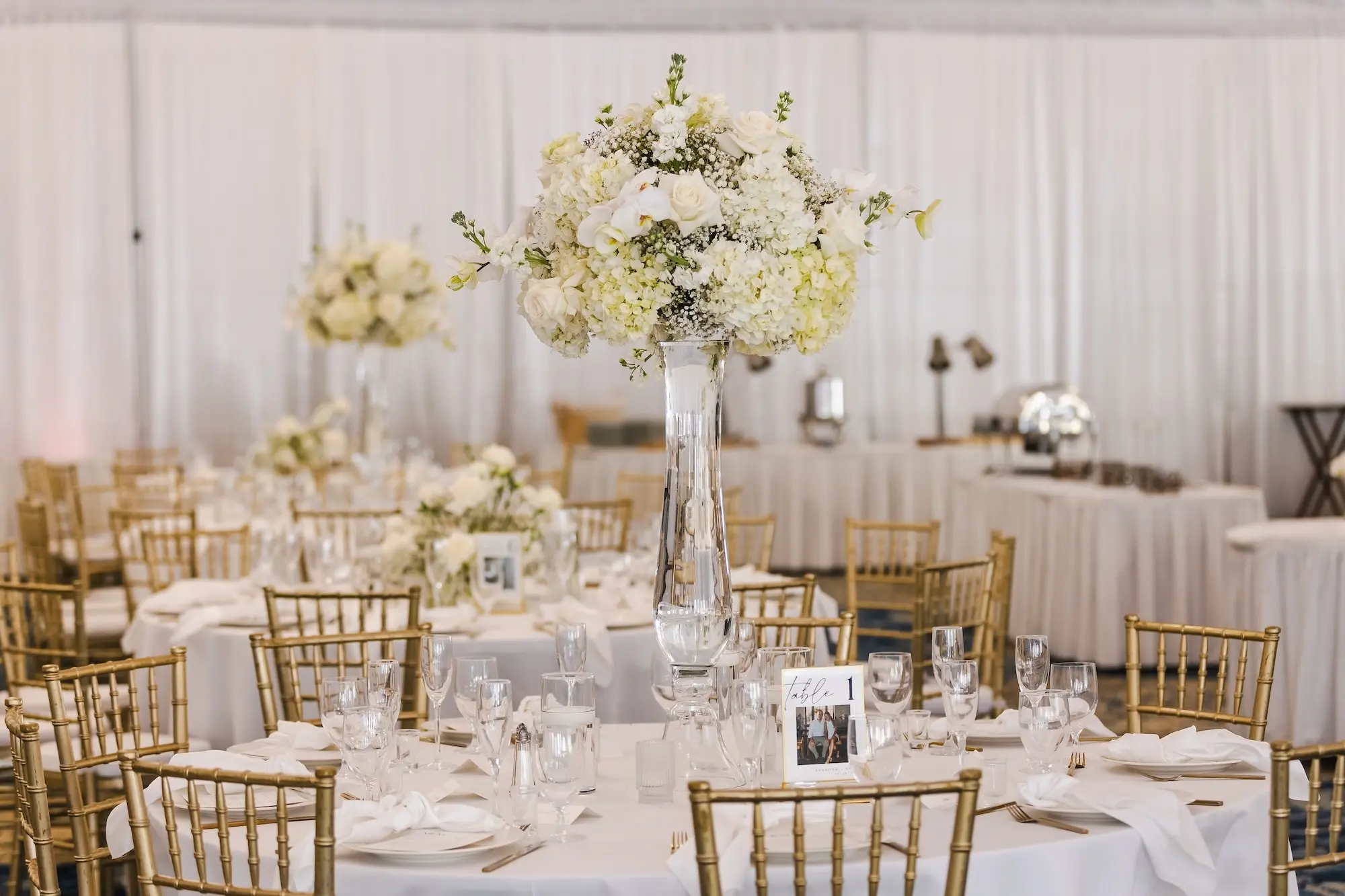 Classic Champagne and Black Florida Beach Tented Ballroom Wedding Reception | White Flower Roses and Baby's Breath in Tall Glass Vase Centerpiece Decor Ideas | Gold Chiavari Chairs at Round Tables with White Linens Decor Inspiration