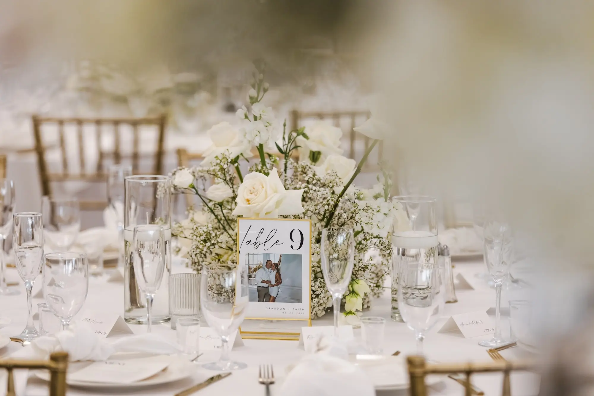 Classic Champagne and Black Florida Beach Tented Ballroom Wedding Reception | White Flower Roses and Baby's Breath Centerpiece Decor Ideas | White Cardstock Pictures with Black Writing in Gold Frame Table Number Inspiration