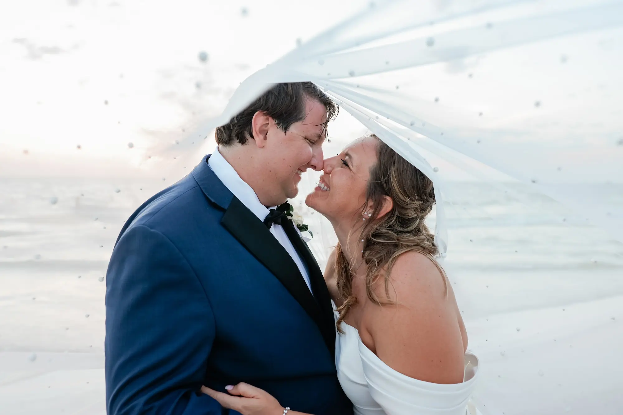 Bride and Groom Waterfront Beach Wedding Portrait Under Veil | Half Up Half Down Beach Curls with Natural Makeup Bridal Look Inspiration | Ybor Hair and Makeup Artist Adore Bridal Hair and Makeup | St Pete Photographer Lifelong Photography Studio