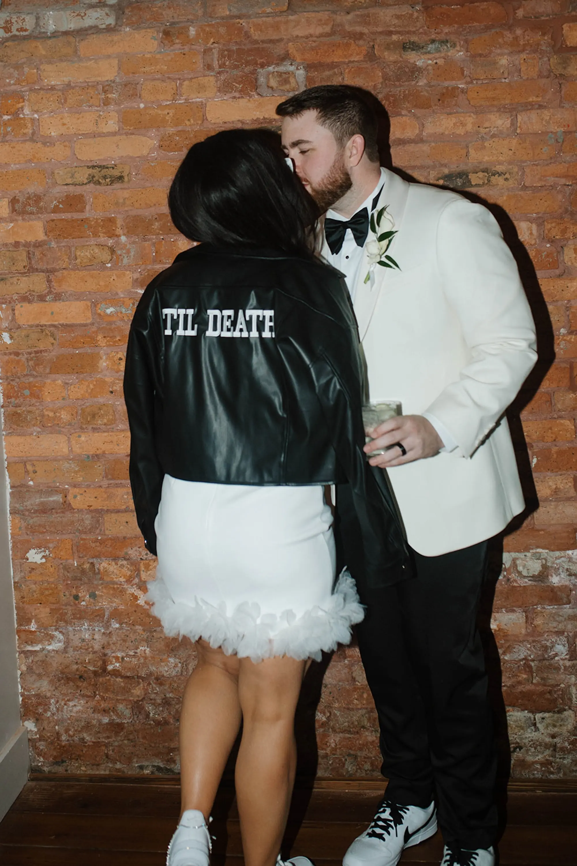 Bride and Groom Wedding Reception Portrait in Front of Industrial Brick Wall | Groom Wedding Portrait | Pearl Off White Groom Wedding Tuxedo Jacket with Black Bow Tie Inspiration | Modern Til Death Personalized Leather Jacket with Reception Wedding Gown Ideas