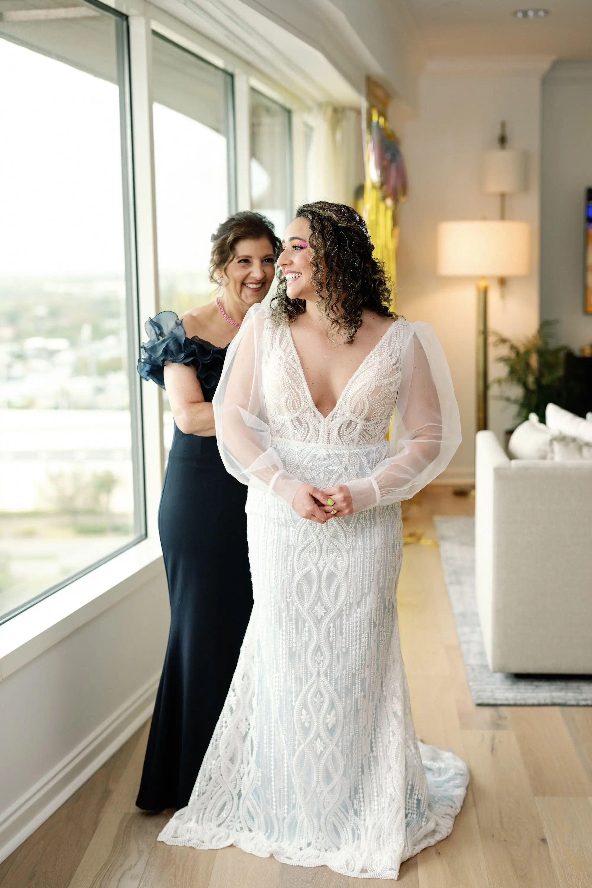 7Whimsical Rainbow Inspired Wedding Ideas - Whimsical, Vibrant Downtown Tampa Wedding | Armature Works Bride and Mother Getting Ready Wedding Portrait | Elegant V-Neck White Magnolia White Wedding Gown with Long Sheer Sleeves Ideas | Half Up Half Down Voluminous Curls Bridal Hair Inspiration | Vibrant Eyeshadow Ideas | Ybor Hair and Makeup Artist Adore Bridal Hair and Makeup | Tampa Bay Photographer Dewitt For Love Photography