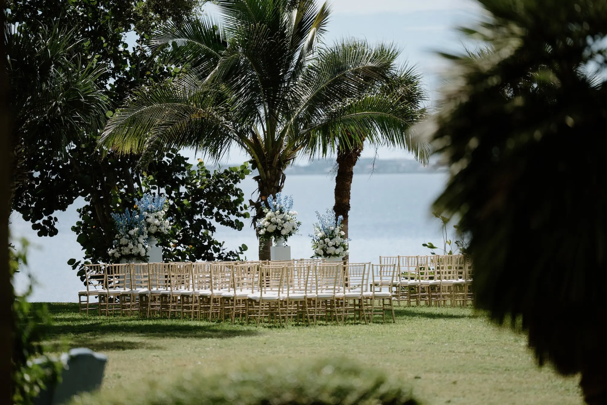 White Roses and Florals with Blue Delphiniums in White Vases on White Pedestals Wedding Ceremony Floral Arrangement Decor Ideas | Ocean View Wedding Reception Inspiration | Gold Chiavari Ceremony Chairs Ideas | Sarasota Florist Beneva Flowers