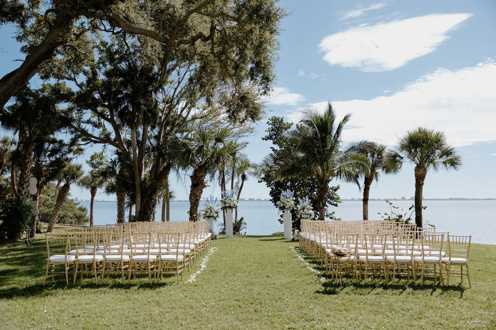 White Roses and Florals with Blue Delphiniums in White Vases on White Pedestals Wedding Ceremony Floral Arrangement Decor Ideas | Ocean View Wedding Reception Inspiration | Gold Chiavari Ceremony Chairs Ideas | Sarasota Florist Beneva Flowers