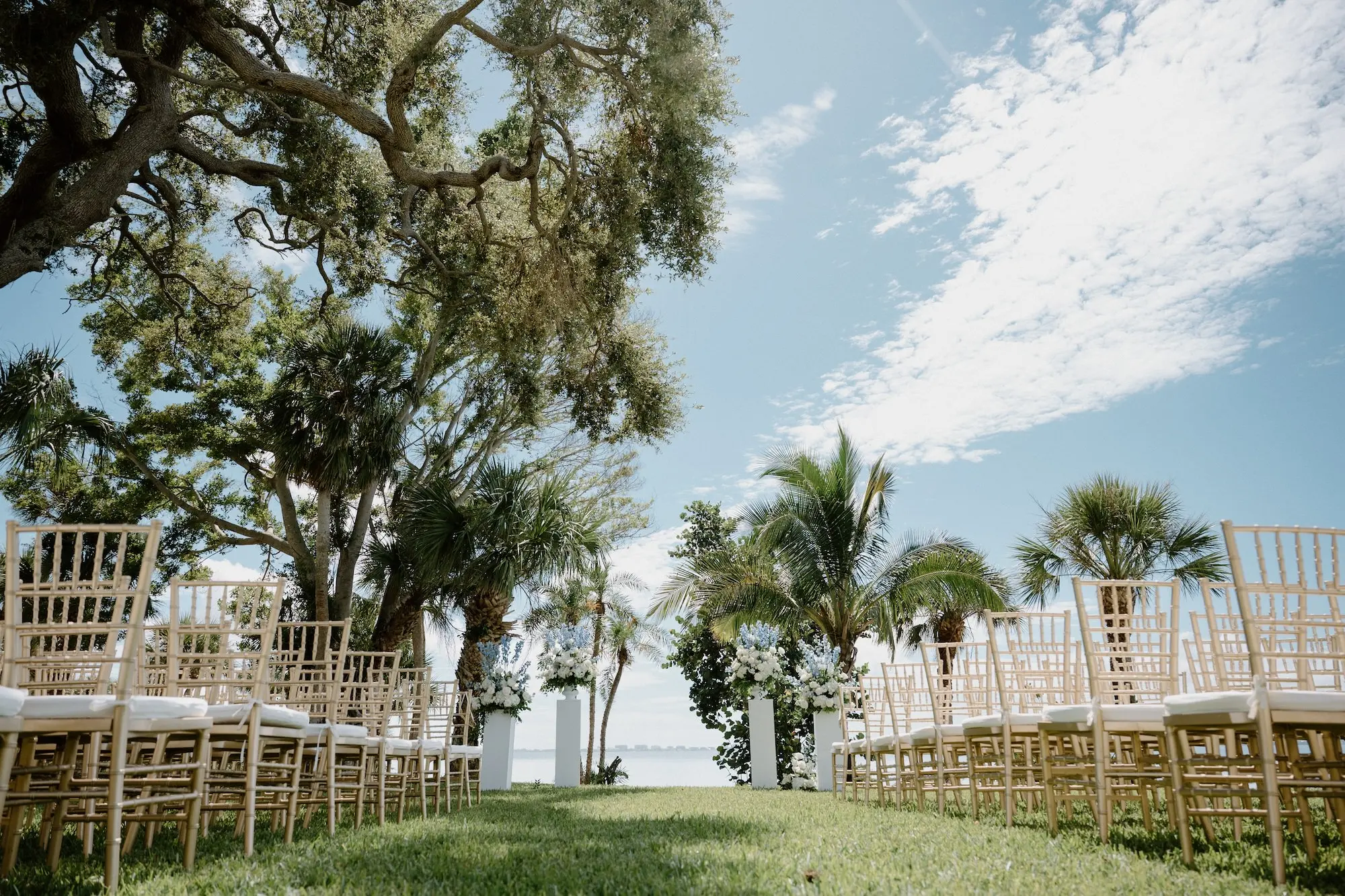 Oceanview Timeless Wedding Reception Venue Inspiration | White Roses and Florals with Blue Delphiniums in White Vases on White Pedestals Wedding Ceremony Floral Arrangement Decor Ideas | Ocean View Wedding Reception Inspiration | Gold Chiavari Ceremony Chairs Ideas | Sarasota Florist Beneva Flowers