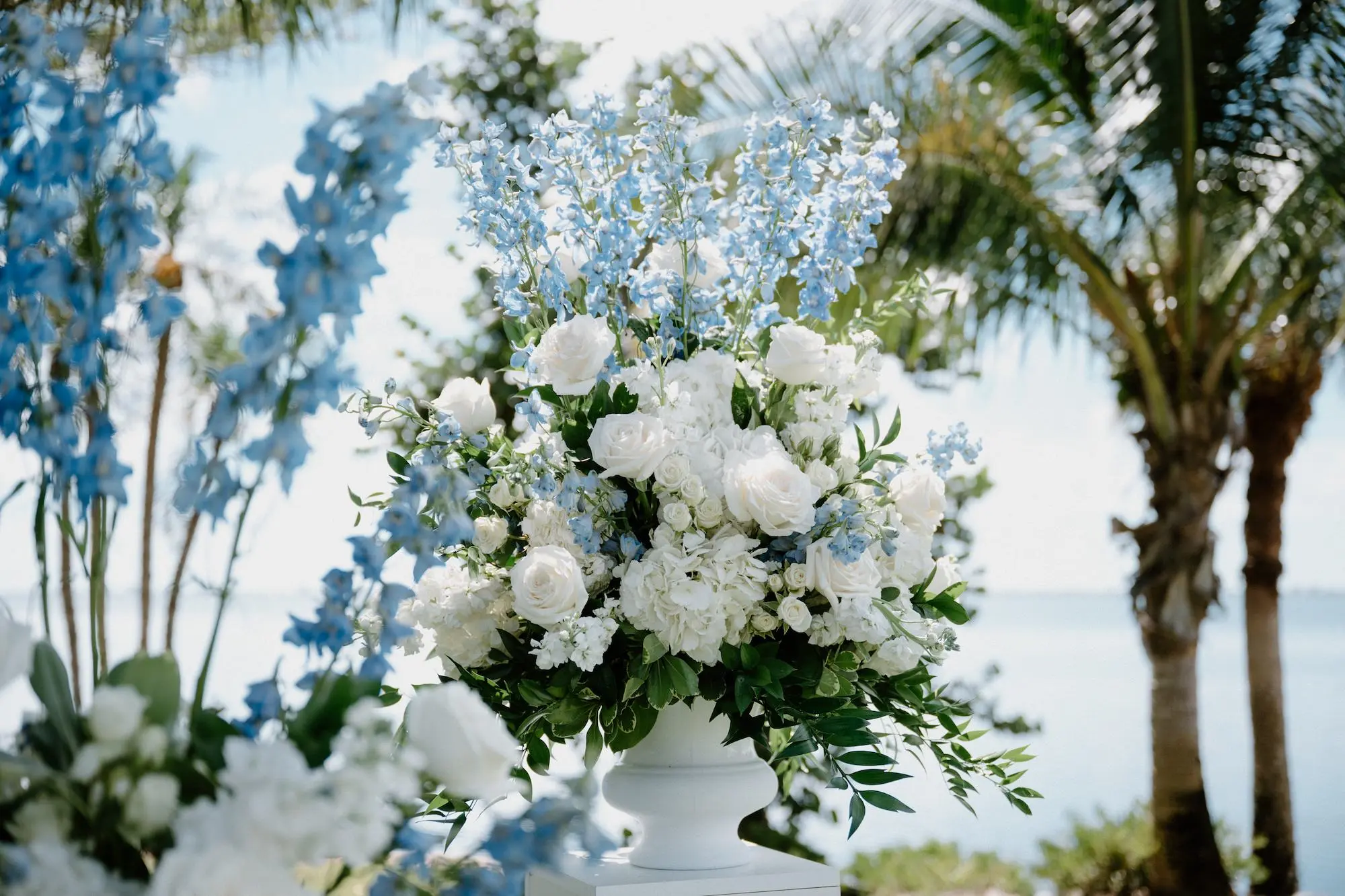 White Roses and Florals with Blue Delphiniums in White Vases on White Pedestals Wedding Ceremony Floral Arrangement Decor Ideas | Ocean View Wedding Reception Inspiration | | Sarasota Florist Beneva Flowers