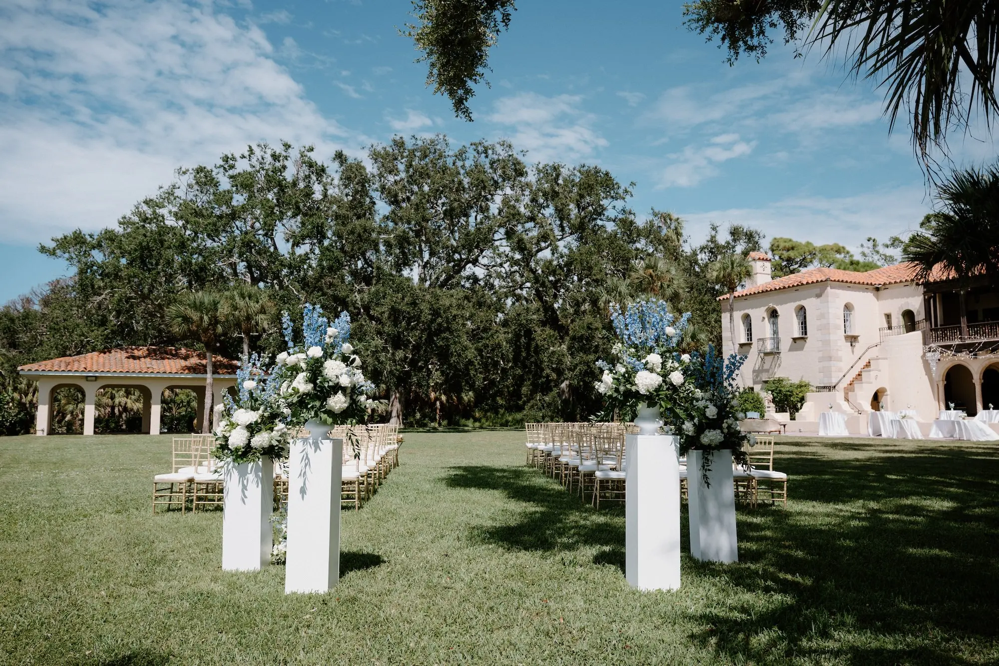 White Roses and Florals with Blue Delphiniums in White Vases on White Pedestals Wedding Ceremony Floral Arrangement Decor Ideas | Ocean View Wedding Reception Inspiration | | Sarasota Florist Beneva Flowers