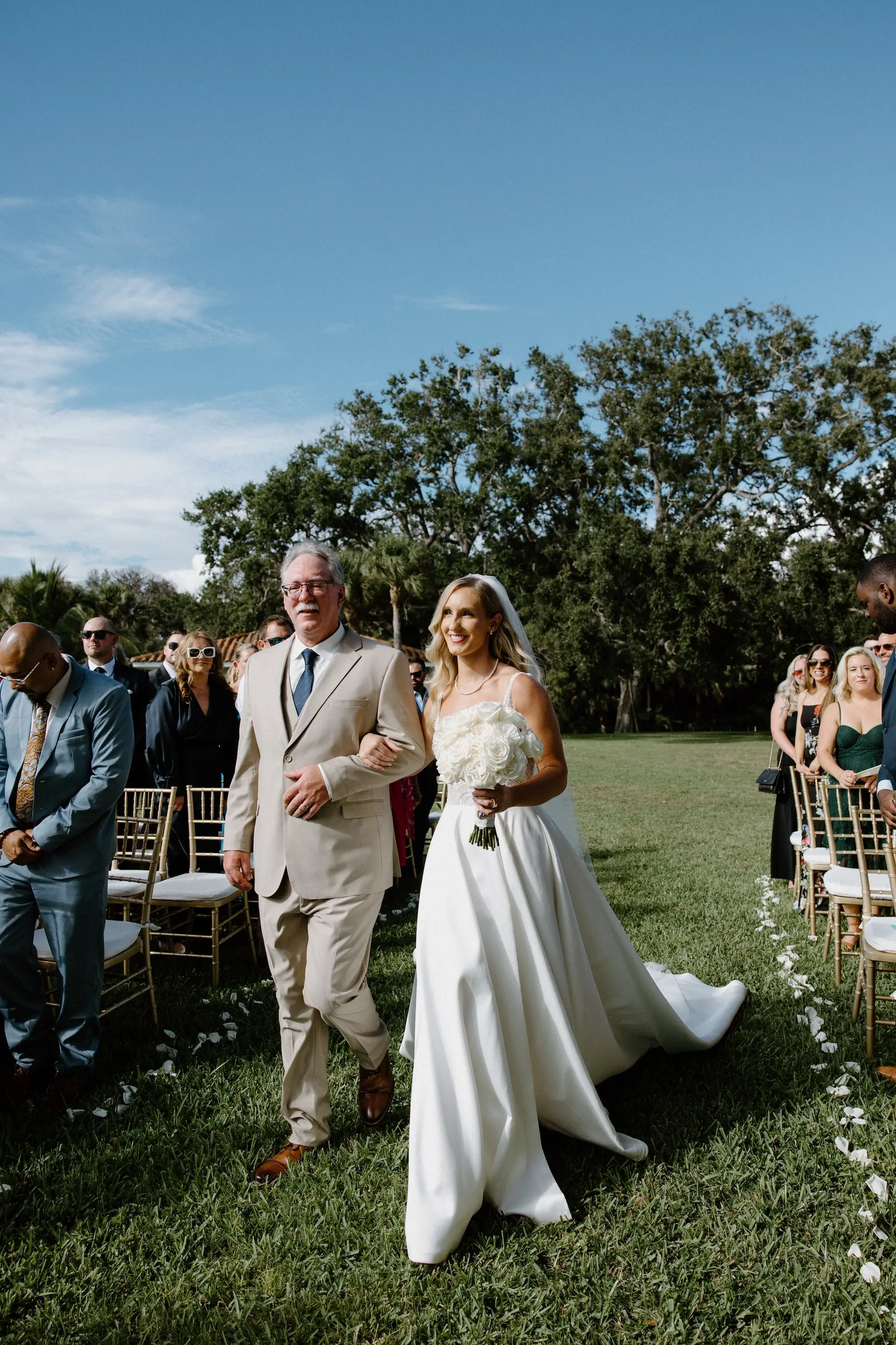 Bride and Father Walking Down Wedding Ceremony Aisle | Monochromatic White Roses Bridal Bouquet Inspiration | Outdoor Estate Wedding Ceremony Ideas | White Pearl Corset Milla Nova Wedding Gown with Dramatic Satin Train Inspiration | All White Groom Wedding Suit Ideas | Monochromatic White Roses Bridal Bouquet Inspiration | Hollywood Glam Curled Waves Wedding Bridal Hair Ideas | Sarasota Florist Beneva Flowers | Tampa Bay Boutique Truly Forever Bridal | Sarasota Hair and Makeup Artist Femme Akoi Beauty Studio