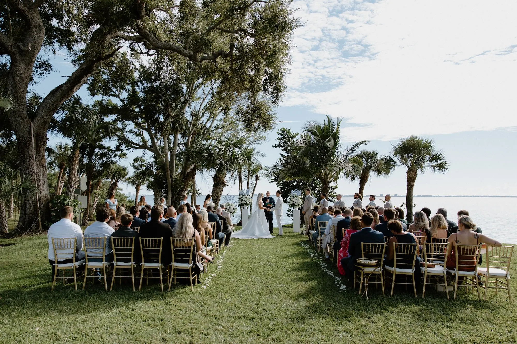 Bride and Groom Wedding Ceremony Vow Exchange Wedding Portrait | White Roses and Florals with Blue Delphiniums in White Vases on White Pedestals Wedding Ceremony Floral Arrangement Decor Ideas | Ocean View Wedding Reception Inspiration | Gold Chiavari Ceremony Chairs Ideas | Sarasota Florist Beneva Flowers